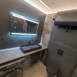 Nowoczesna łazienka z czarnymi płytkami, podświetlanym lustrem, umywalką nablatową i toaletą w minimalistycznym stylu, z widocznym bojlerem elektrycznym na ścianie.