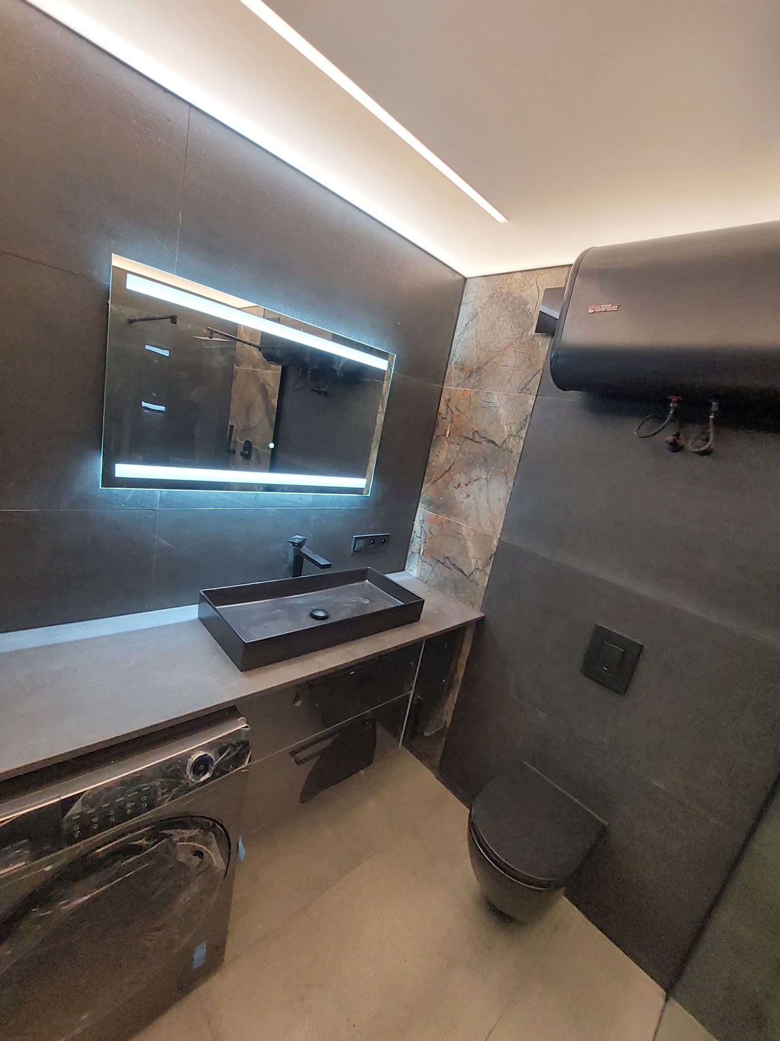 Nowoczesna łazienka z czarnymi płytkami, podświetlanym lustrem, umywalką nablatową i toaletą w minimalistycznym stylu, z widocznym bojlerem elektrycznym na ścianie.