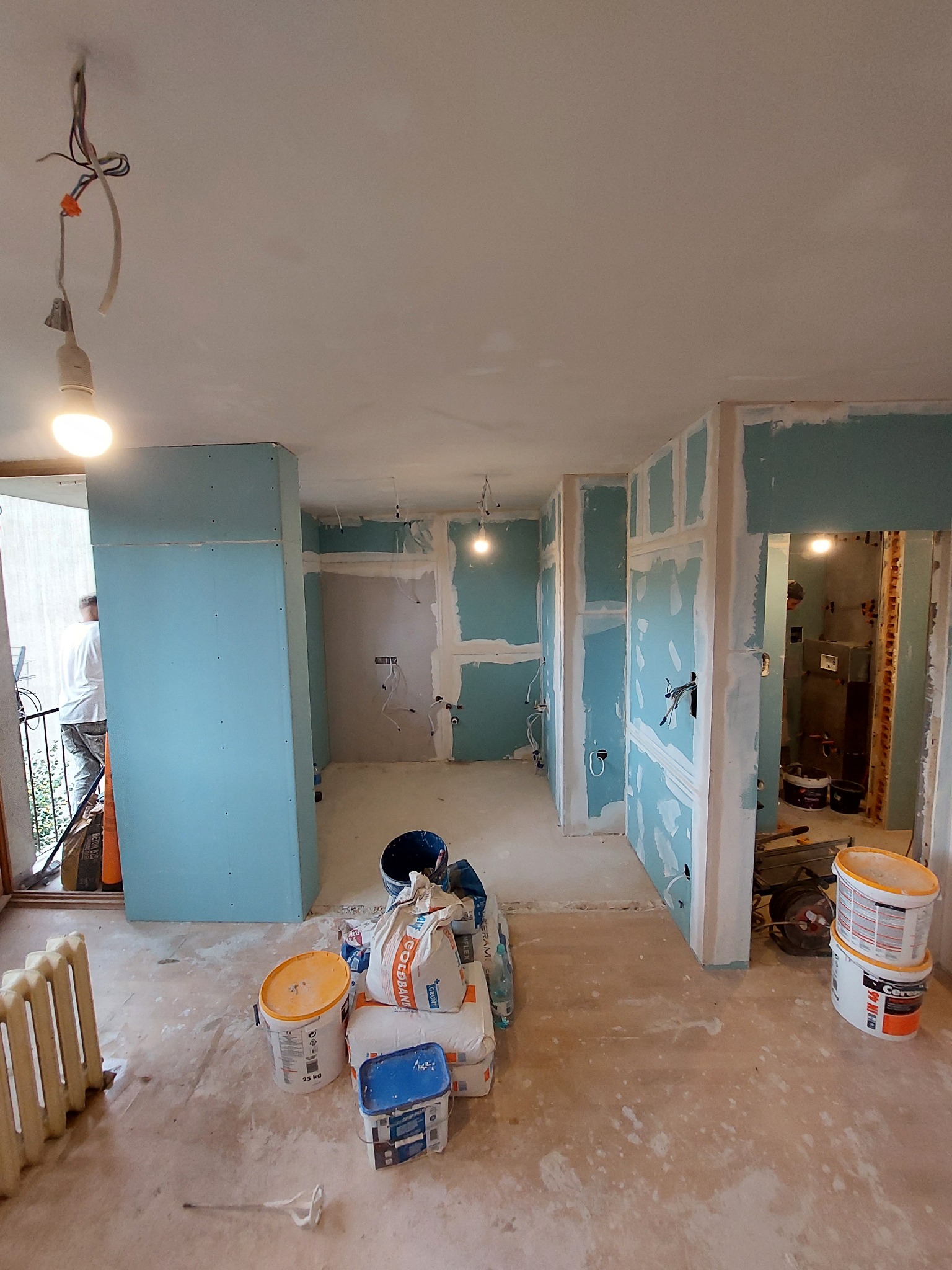 Wnętrze mieszkania w trakcie prac wykończeniowych: ściany pokryte płytami gipsowo-kartonowymi, widoczne puszki elektryczne, worki z zaprawą i wiadra z farbą na podłodze, tymczasowe oświetlenie...