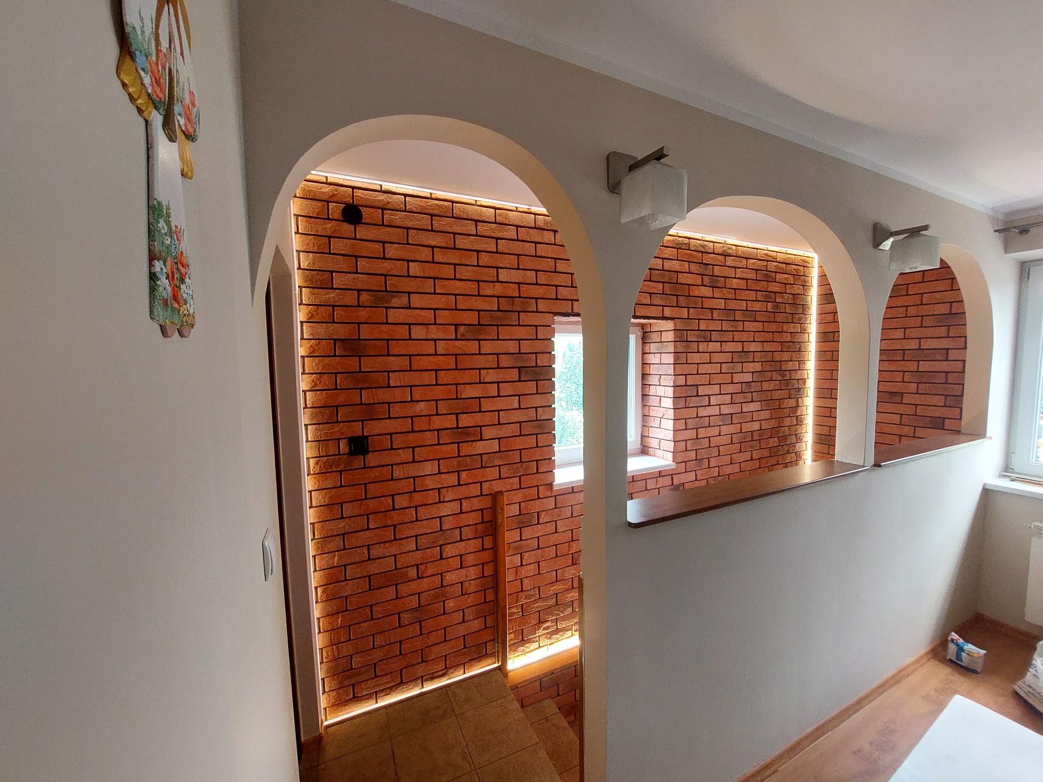 Aranżacja wnętrza z trzema łukowatymi przejściami, ściana wyłożona czerwoną cegłą z podświetleniem LED, widoczne schody i dekoracja ścienna.