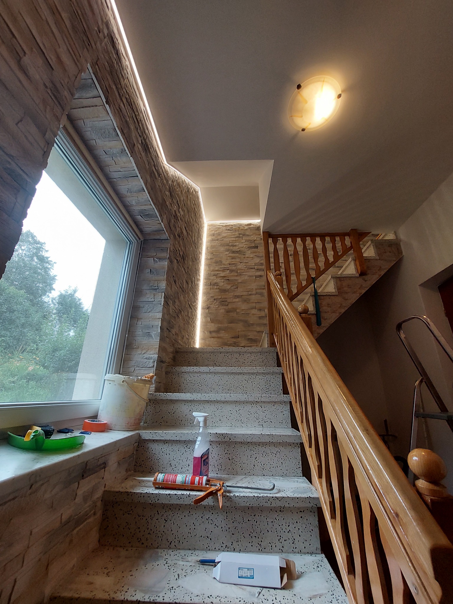 Klatka schodowa w trakcie wykańczania z oknem, obłożona kamieniem dekoracyjnym z ledowym oświetleniem, widoczne narzędzia i materiały budowlane.