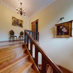 Wnętrze z drewnianymi schodami z rzeźbioną balustradą, kolumną dekoracyjną, dwoma obrazami w złotych ramach i ozdobnym żyrandolem.