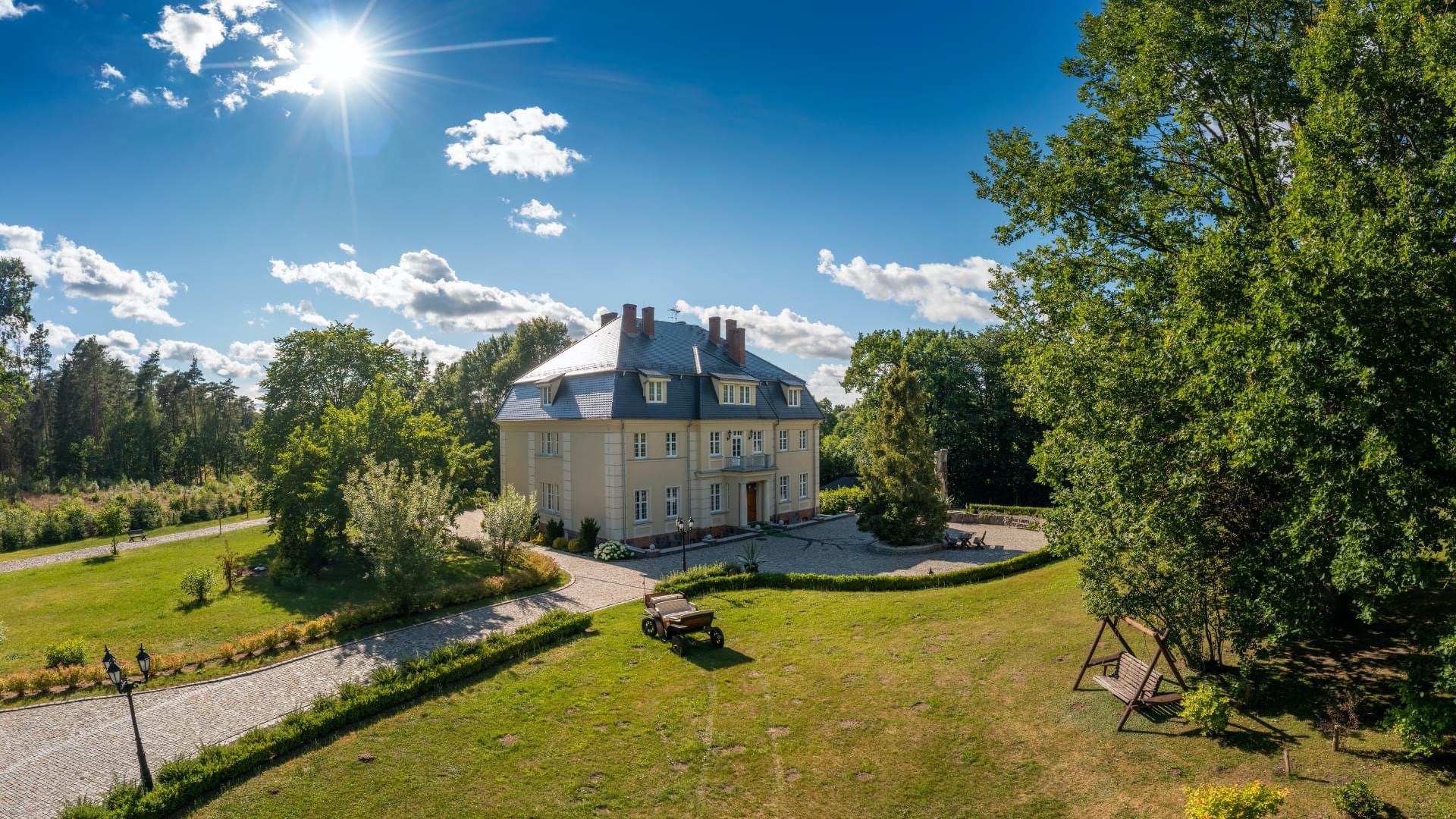 Elegancki, dwukondygnacyjny budynek z szarym dachem, otoczony zielenią, z wozem na trawniku i huśtawką w cieniu drzew, widok z góry w słoneczny dzień.