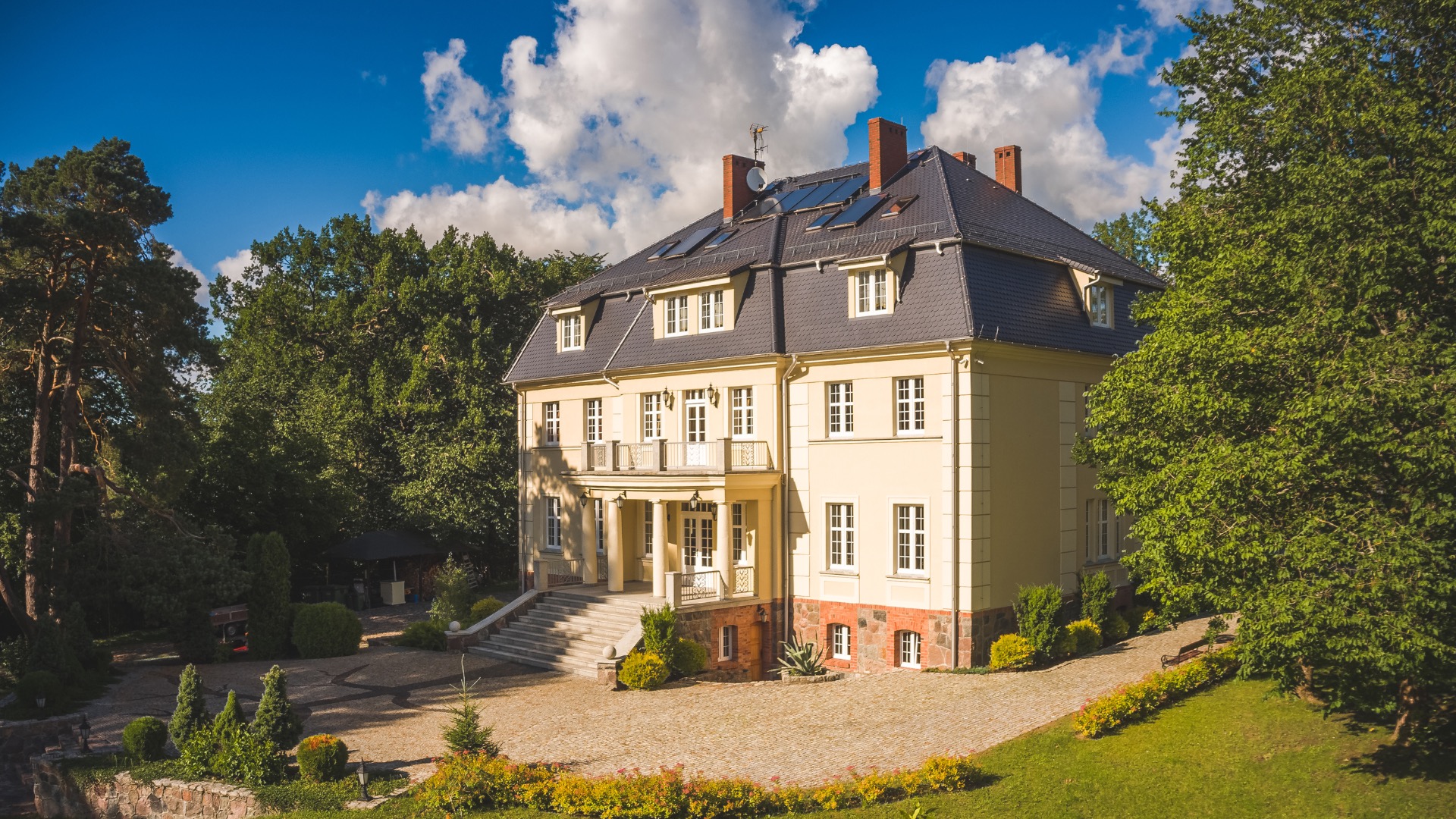 Elegancki, trzypiętrowy budynek kliniki w Słupsku z kolumnami, balkonami i panelami słonecznymi na dachu, otoczony zielenią i brukowanym podjazdem.
