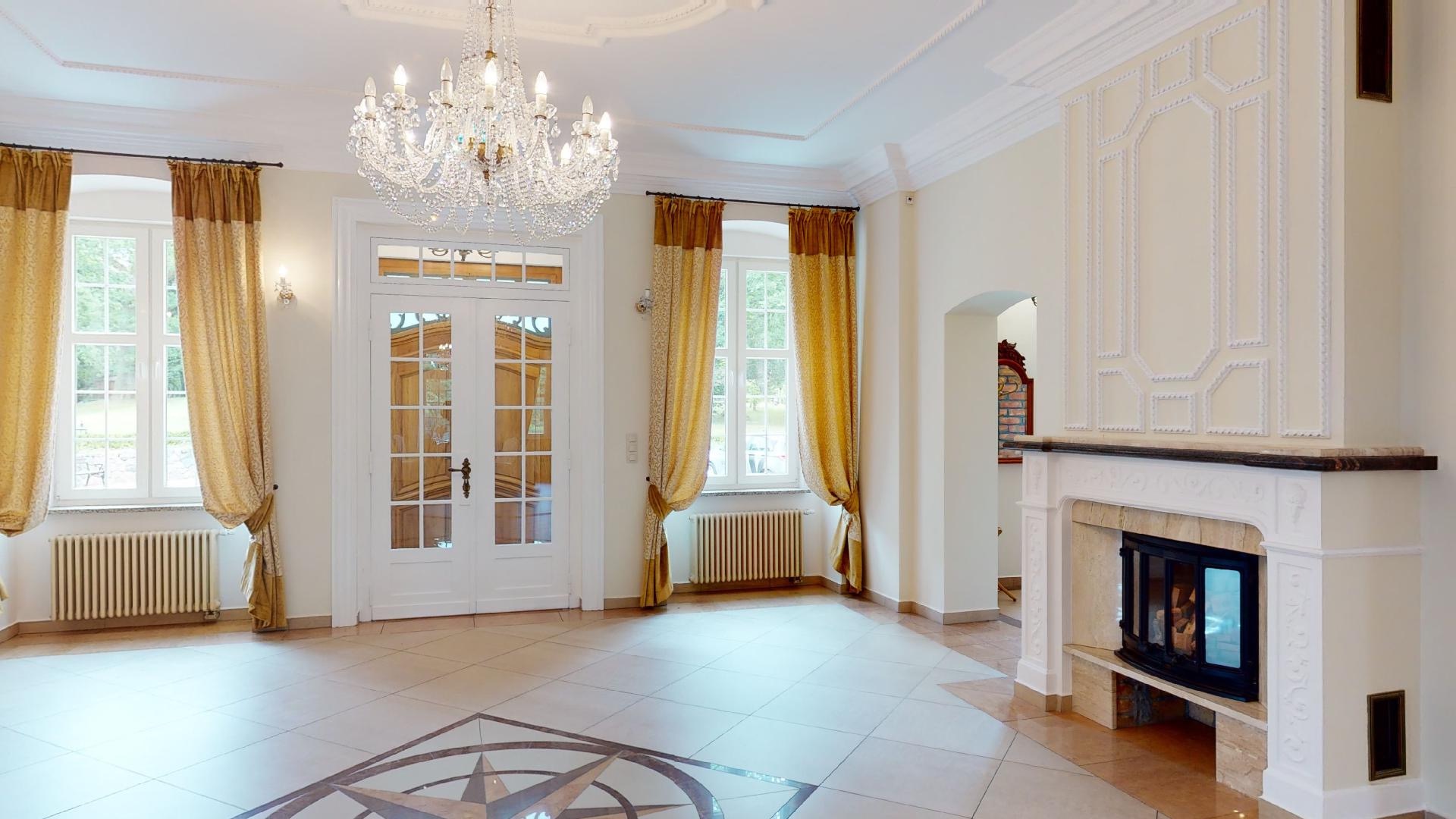 Eleganckie wnętrze z kryształowym żyrandolem, białymi drzwiami z przeszkleniami, oknami z zasłonami w kolorze złota i kominkiem z dekoracyjną obudową.