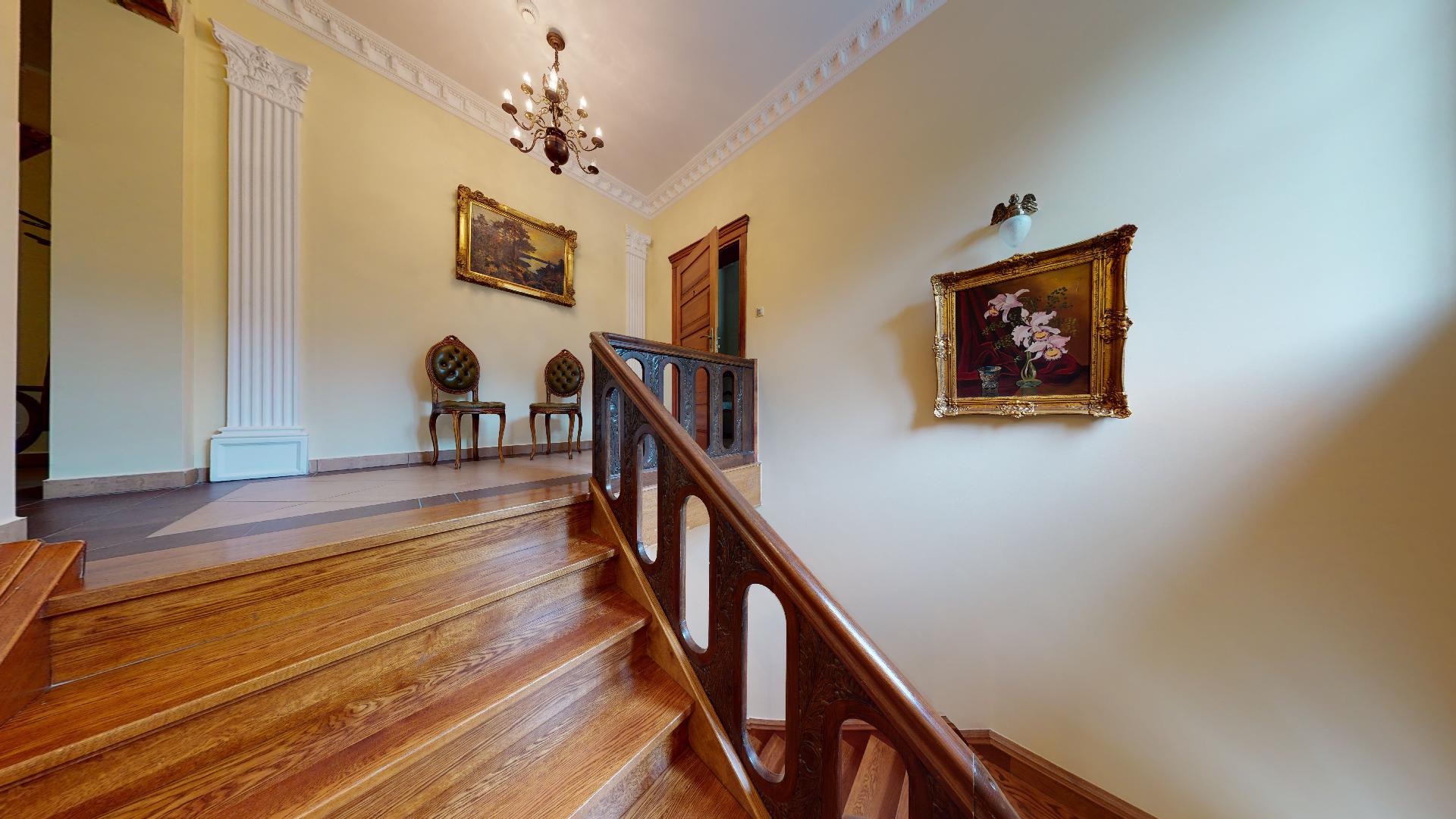 Wnętrze z drewnianymi schodami z rzeźbioną balustradą, kolumną dekoracyjną, dwoma obrazami w złotych ramach i ozdobnym żyrandolem.