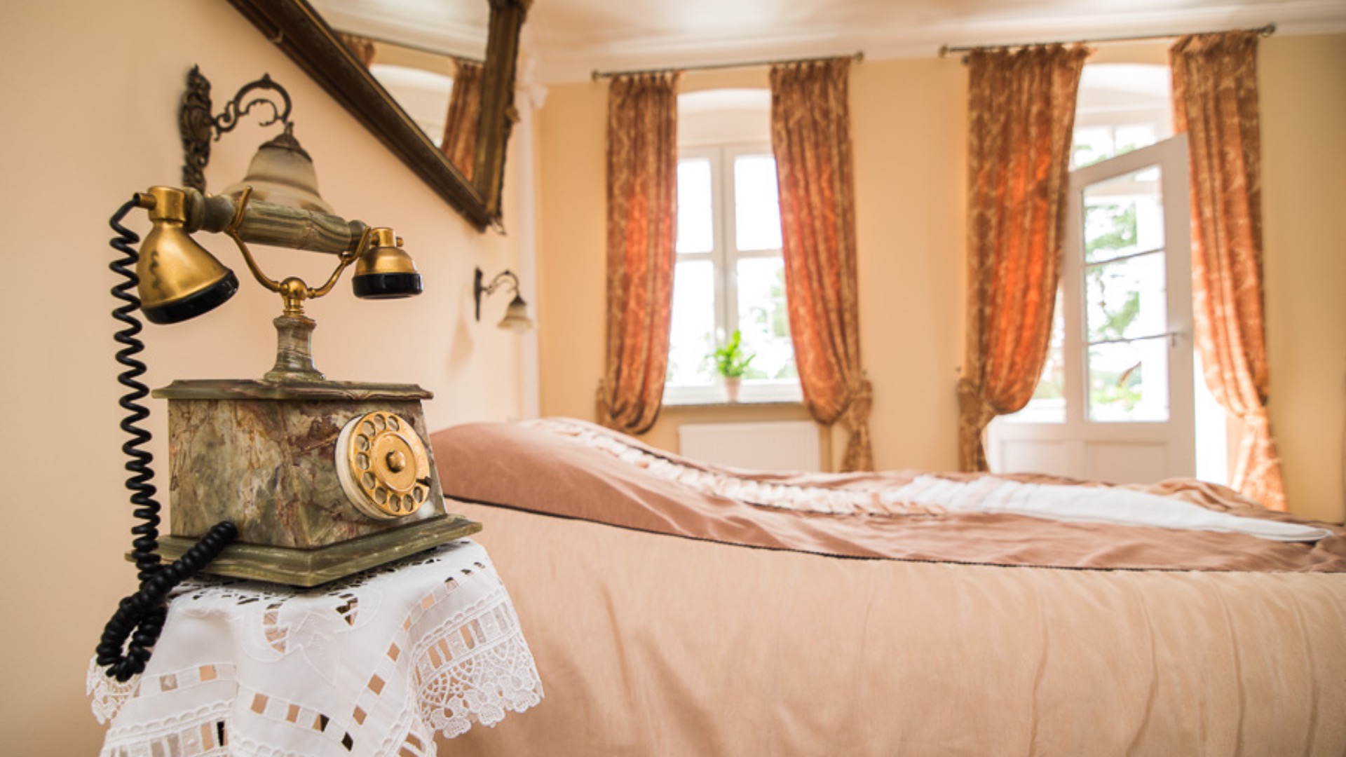 Stylowy pokój z łóżkiem i zabytkowym telefonem na stoliku nocnym, okna z ozdobnymi zasłonami w tle.