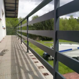 Grafitowa poręcz balkonowa z metalowych profili na tle zielonego ogrodu, widoczny fragment białego samochodu dostawczego