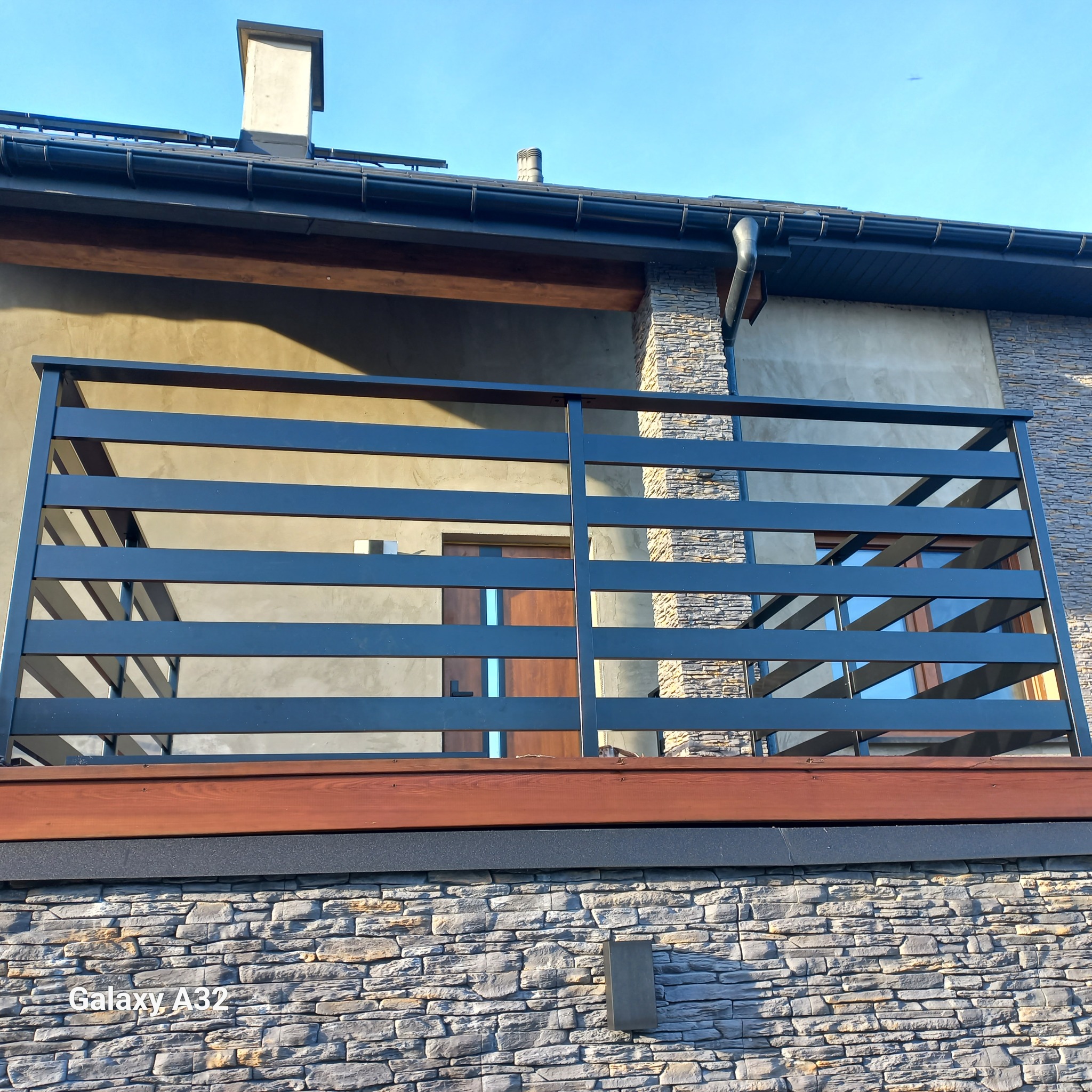 Nowoczesna, pozioma, metalowa balustrada balkonowa w kolorze grafitowym, zamontowana na balkonie budynku z elewacją z kamienia łupanego. Widoczne drewniane elementy wykończeniowe.