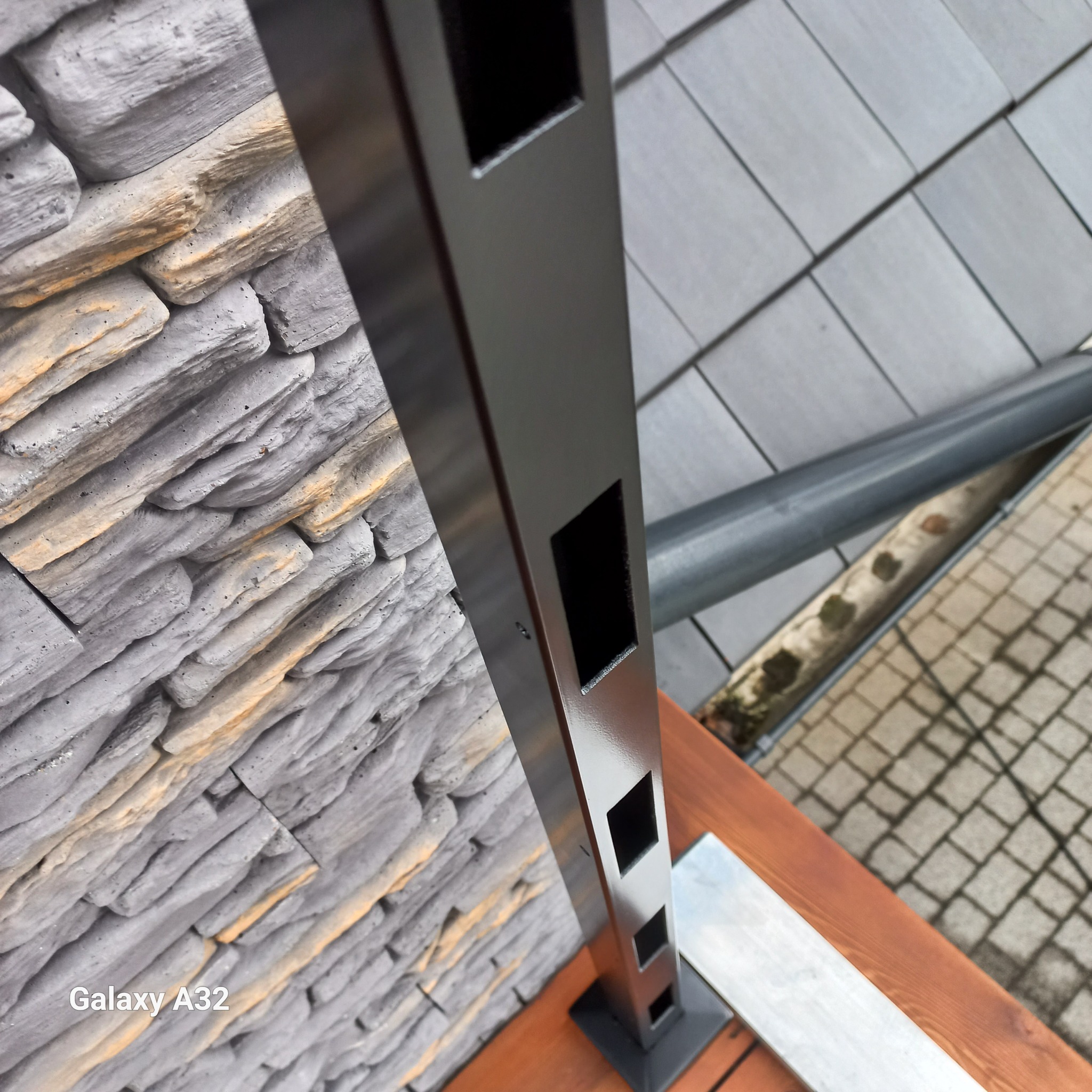 Metalowy słupek balustrady z kwadratowymi otworami, zamontowany na drewnianym tarasie, na tle kamiennej ściany i szarej nawierzchni.
