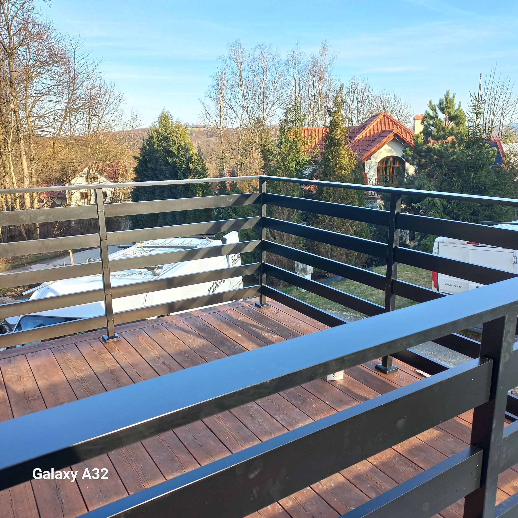 Nowoczesna, czarna balustrada z poziomych profili na drewnianym tarasie, z widokiem na okoliczne domy i wzgórza w słoneczny dzień.