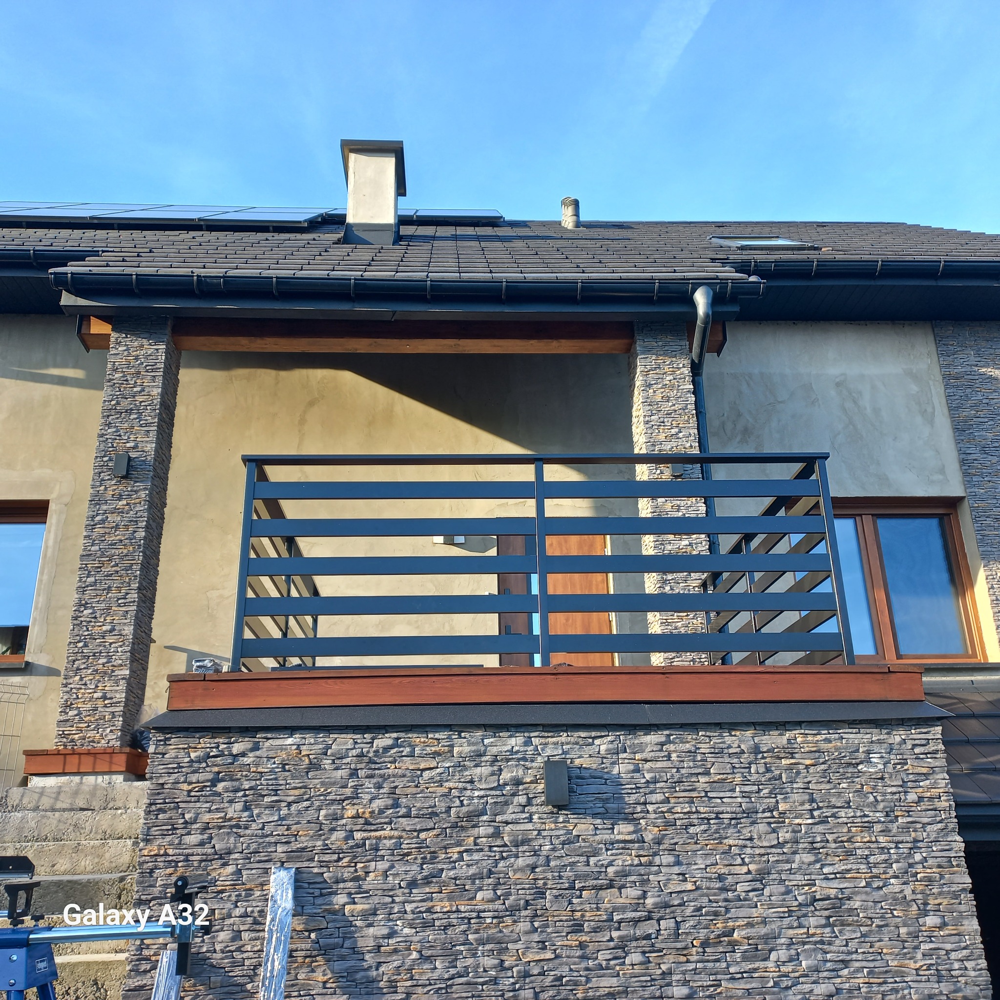 Nowoczesna, pozioma balustrada balkonowa w kolorze grafitowym, zamontowana na balkonie domu z elewacją wykończoną kamieniem dekoracyjnym.