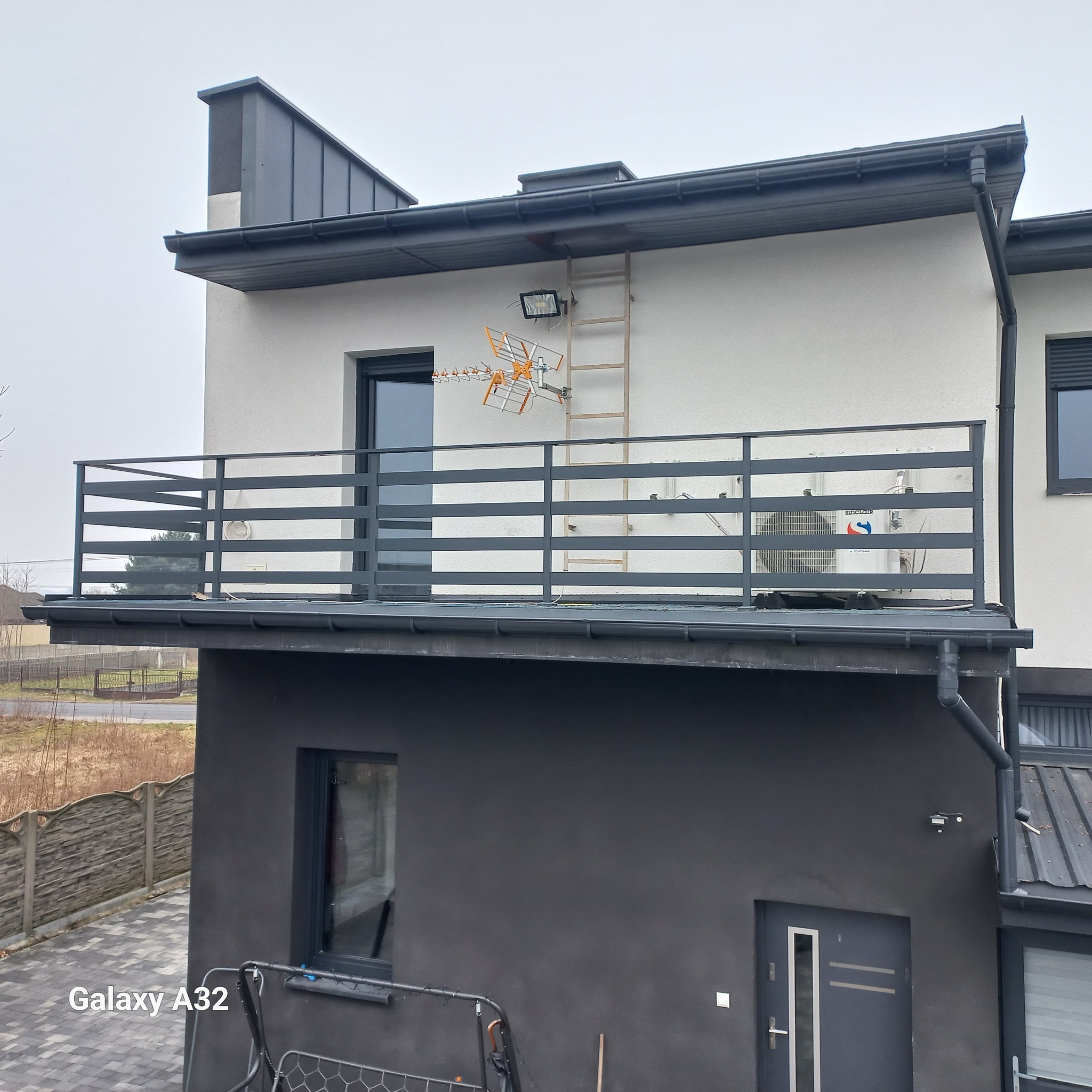 Nowoczesny balkon z szarą, poziomą balustradą na tle dwukondygnacyjnego domu z anteną i drabinką na ścianie.
