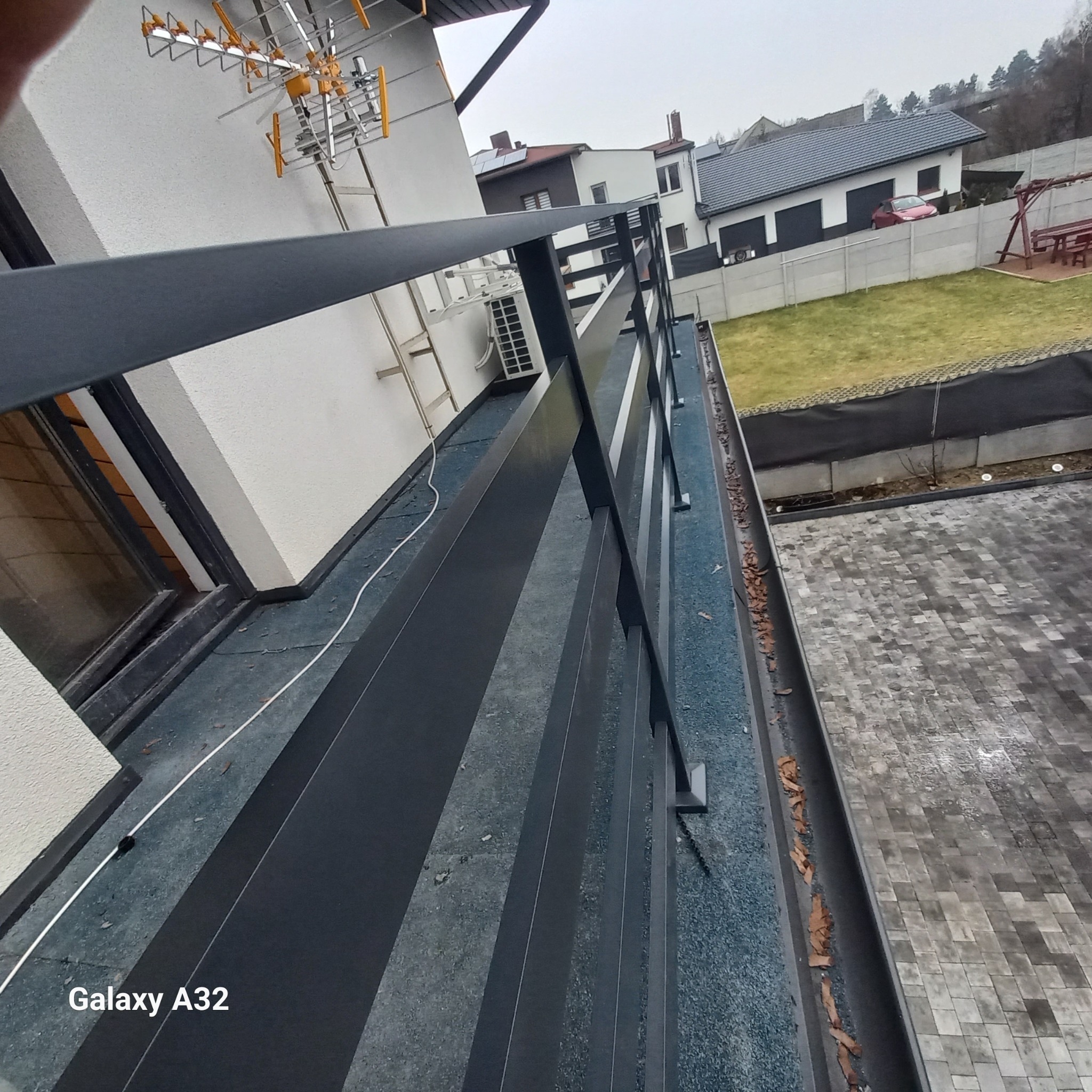 Nowoczesna, ciemnoszara balustrada balkonowa z metalowymi słupkami na tle domu jednorodzinnego z anteną na dachu i widokiem na ogród z betonowym ogrodzeniem.