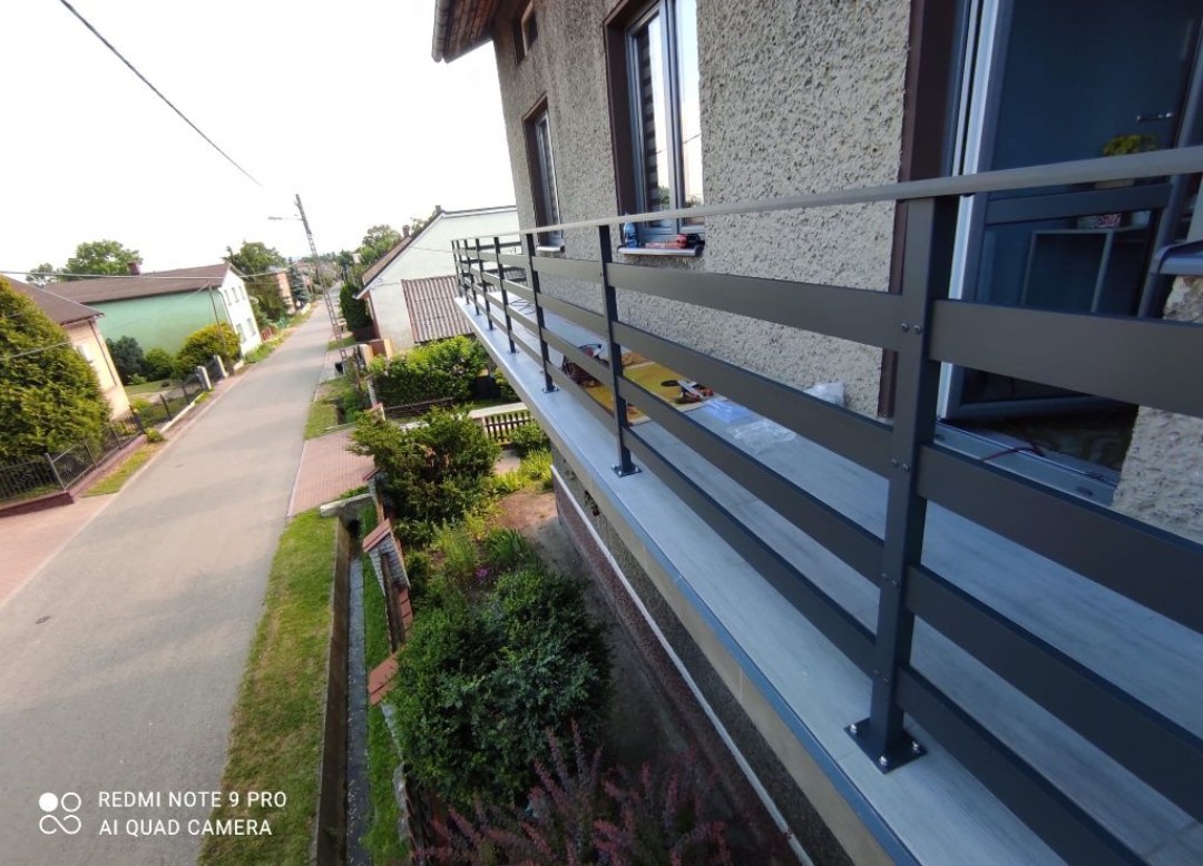 Nowoczesna, grafitowa balustrada balkonowa z poziomymi szczeblami, zamontowana przy budynku z elewacją w odcieniach beżu, widok z góry na ulicę i zieleń.