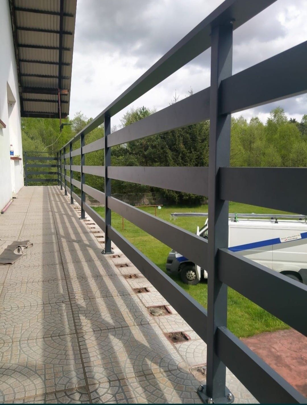 Grafitowa poręcz balkonowa z metalowych profili na tle zielonego ogrodu, widoczny fragment białego samochodu dostawczego