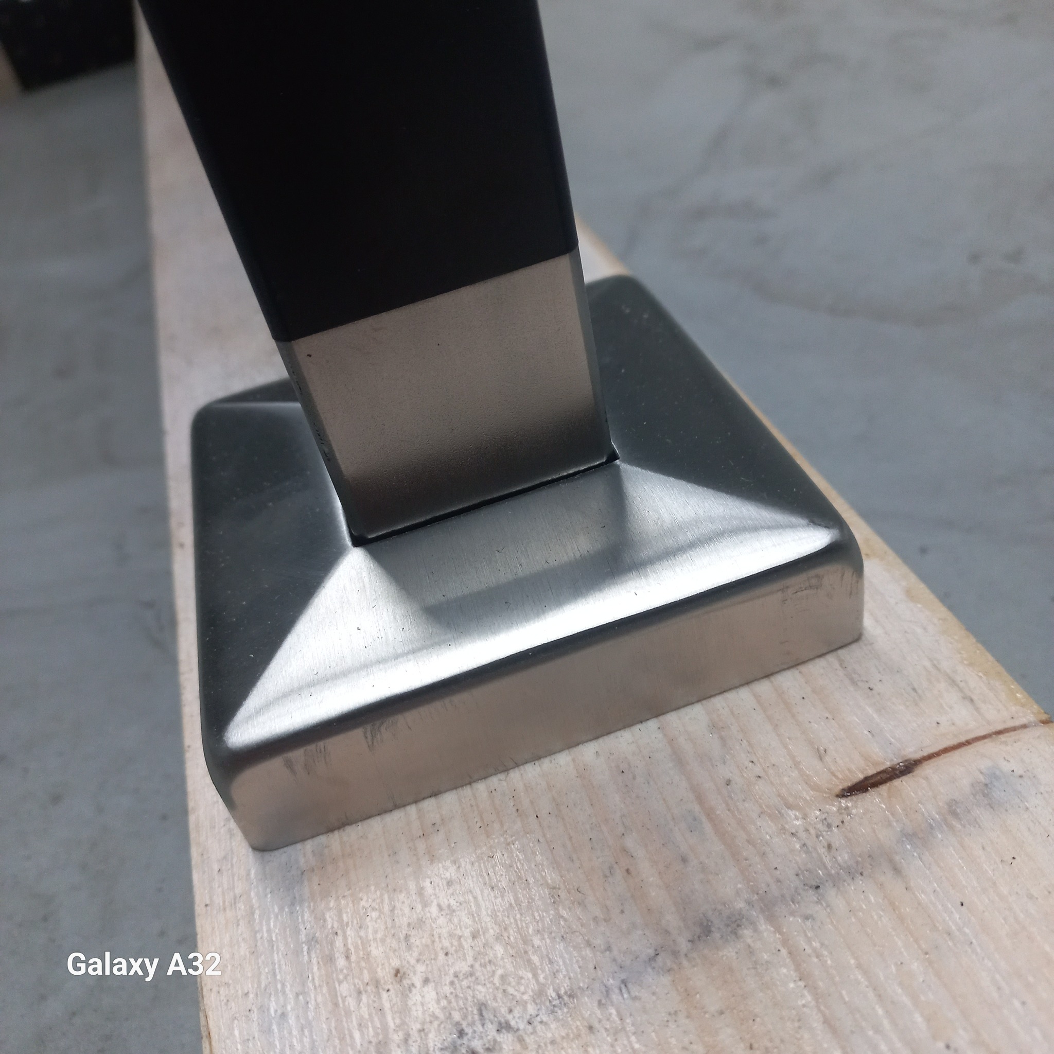 Metalowa podstawa słupka, prawdopodobnie elementu ogrodzenia lub balustrady, zamontowana na drewnianej powierzchni, widoczne ślady obróbki metalu.