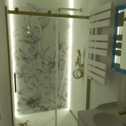 Prywatna - Nowoczesna kabina prysznicowa ze szklanymi drzwiami przesuwnymi z motywem kwiatowym, podświetlona od wewnątrz, obok biały grzejnik drabinkowy i umywalka z baterią w kolorze złotym.