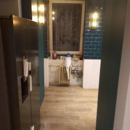 Prywatna - Wnętrze w trakcie prac: widok na podłogę z płytek imitujących drewno, fragment ściany z ciemnozielonej glazury i grzejnik pod oknem, widoczne elementy instalacji wodnej, w tle fragment czarnej...