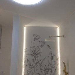 Prywatna - Biała łazienka w Nowym Dworze Mazowieckim z motywem kwiatowym na ścianie, podświetlona listwą LED, okrągła lampa sufitowa i złota słuchawka prysznicowa.