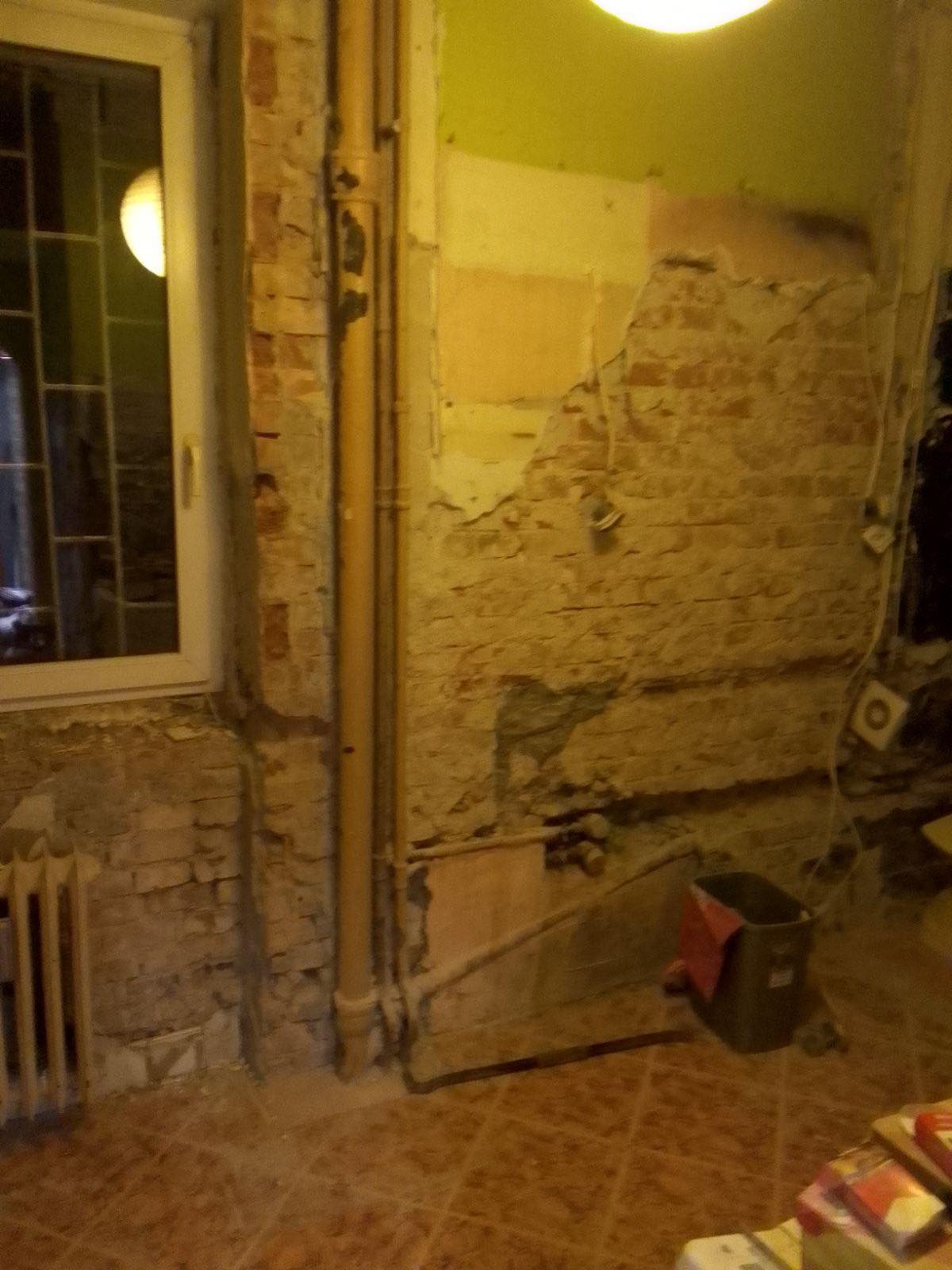 Odkryta cegła na ścianie w trakcie prac remontowych, widoczne instalacje, rury, fragment okna i grzejnika, podłoga wyłożona brązowymi płytkami, w tle wiadro i materiały budowlane.