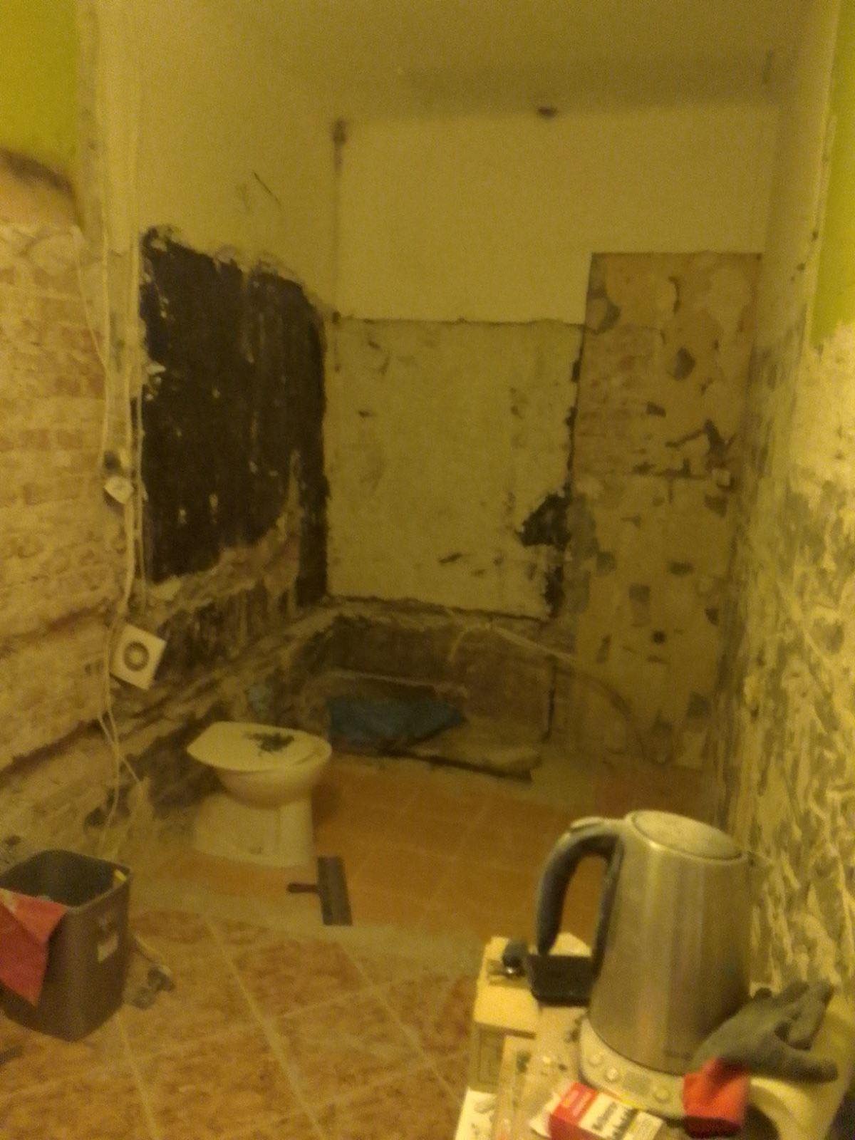 Łazienka w trakcie demontażu: ściany częściowo pozbawione płytek, widoczna instalacja elektryczna, toaleta i narzędzia na podłodze, czajnik elektryczny na blacie.
