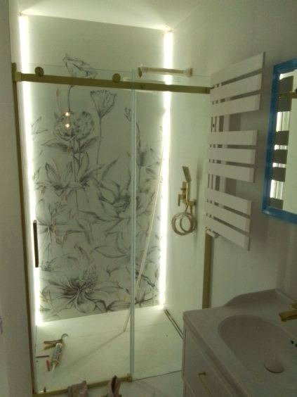 Nowoczesna kabina prysznicowa ze szklanymi drzwiami przesuwnymi z motywem kwiatowym, podświetlona od wewnątrz, obok biały grzejnik drabinkowy i umywalka z baterią w kolorze złotym.