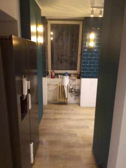 Wnętrze w trakcie prac: widok na podłogę z płytek imitujących drewno, fragment ściany z ciemnozielonej glazury i grzejnik pod oknem, widoczne elementy instalacji wodnej, w tle fragment czarnej...