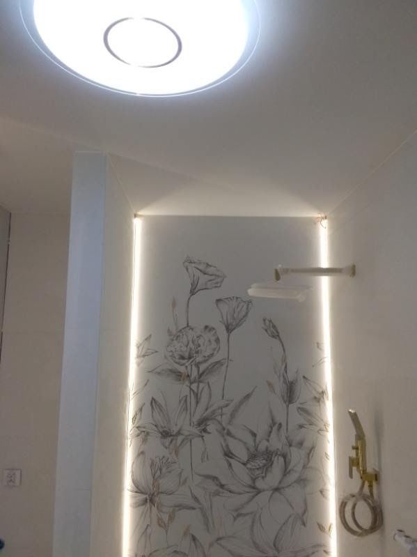 Biała łazienka w Nowym Dworze Mazowieckim z motywem kwiatowym na ścianie, podświetlona listwą LED, okrągła lampa sufitowa i złota słuchawka prysznicowa.