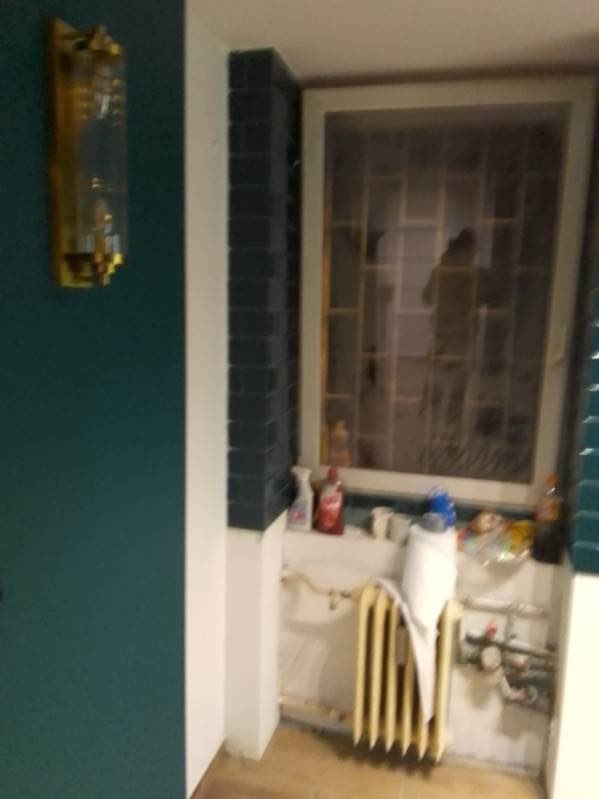 Narożnik pokoju po remoncie z pomalowaną na ciemnozielono ścianą, ozdobną lampą na ścianie, obudowanym kolumną z ciemnych płytek oknem i grzejnikiem pod oknem.