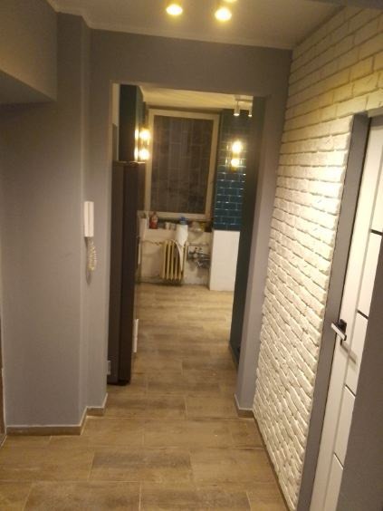 Korytarz po remoncie z beżową podłogą, szarą ścianą i ścianą z białej cegły dekoracyjnej. W głębi widoczny pokój z grzejnikiem i niebieskimi płytkami.