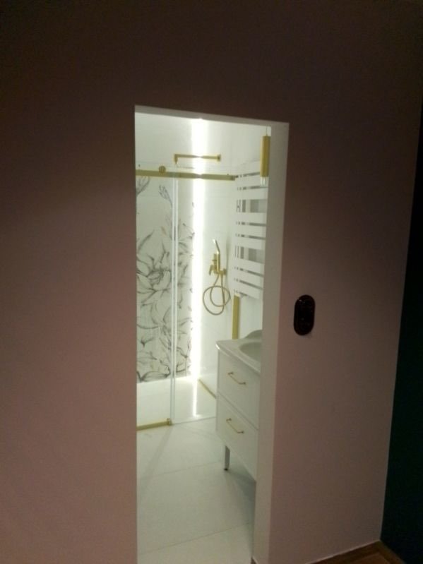 Widok na wykończoną łazienkę z kabiną prysznicową ze złotymi elementami, umywalką z szafką i grzejnikiem drabinkowym, widzianą z korytarza przez otwór drzwiowy w ścianie pomalowanej na różowo.