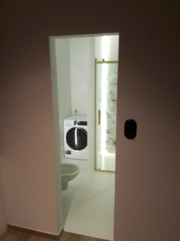 Wnętrze łazienki z widoczną pralką, toaletą i kabiną prysznicową z zasłoną. Ściany pomalowane na biało, widoczny fragment ściany w kolorze pudrowego różu z czarnym włącznikiem światła.
