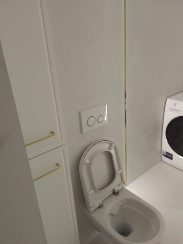 Biała toaleta z otwartą klapą w nowoczesnej łazience z białymi płytkami i szafką z złotymi uchwytami, obok pralka.