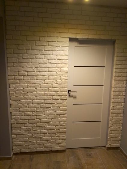 Wykończenie ściany imitacją cegły w odcieniach beżu, sąsiadujące z białą, nowoczesną wewnętrzną stolarką drzwiową z poziomymi frezowaniami.