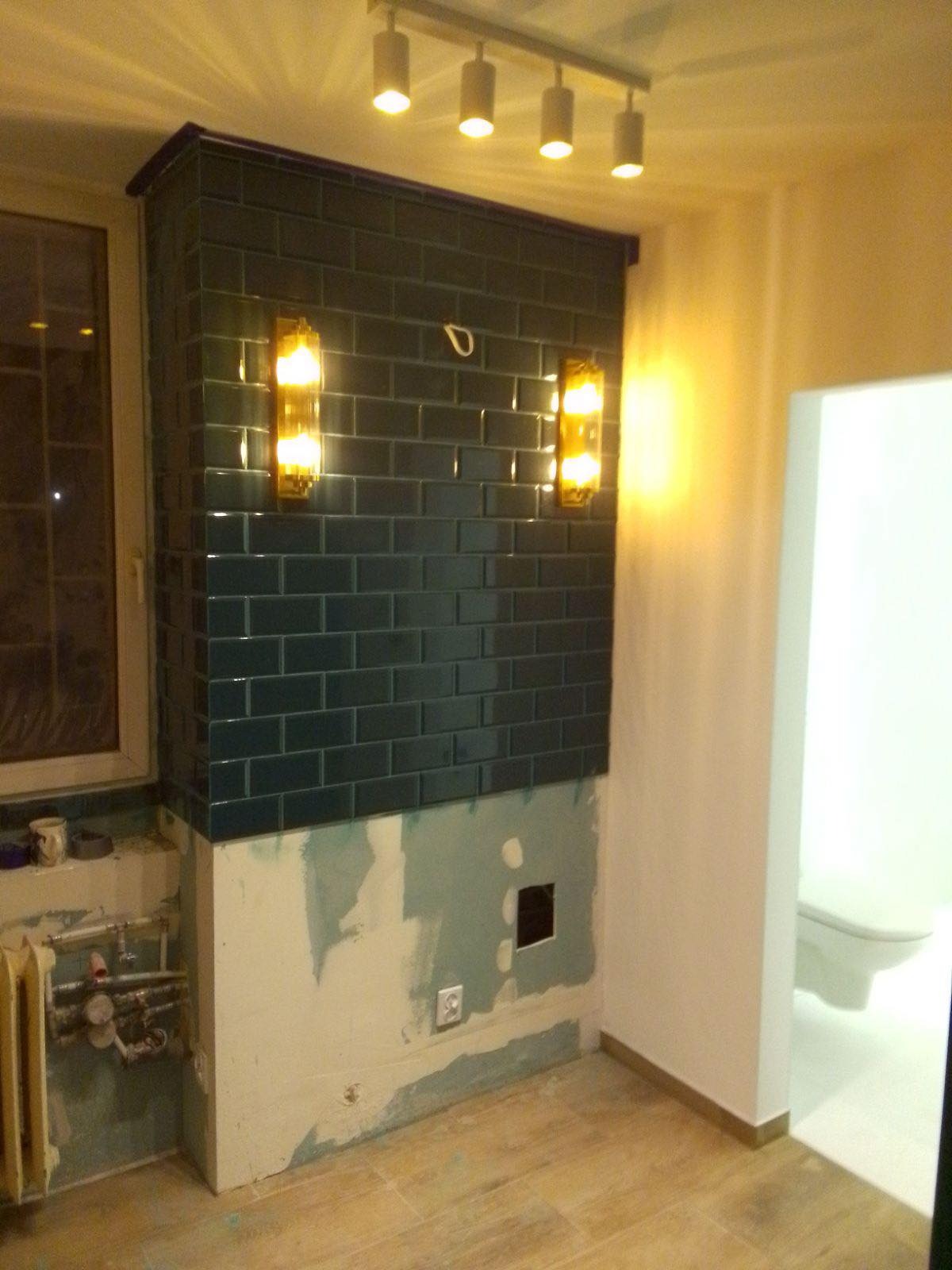 Wnętrze łazienki w trakcie remontu: ściana wyłożona ciemnymi, błyszczącymi płytkami w stylu metro, z zamontowanymi kinkietami, widoczne niedokończone prace wykończeniowe na ścianach i podłodze...