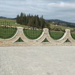 Kamieniarstwo Kacper Wójcik - Kamienny murek z kutymi elementami ogrodzenia, widok na zielone wzgórza w tle, oraz ułożona kostka brukowa na podjeździe.