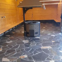 Kamienna posadzka z nieregularnych płyt w altanie z drewnianą zabudową i centralnie umieszczonym paleniskiem z kominem.