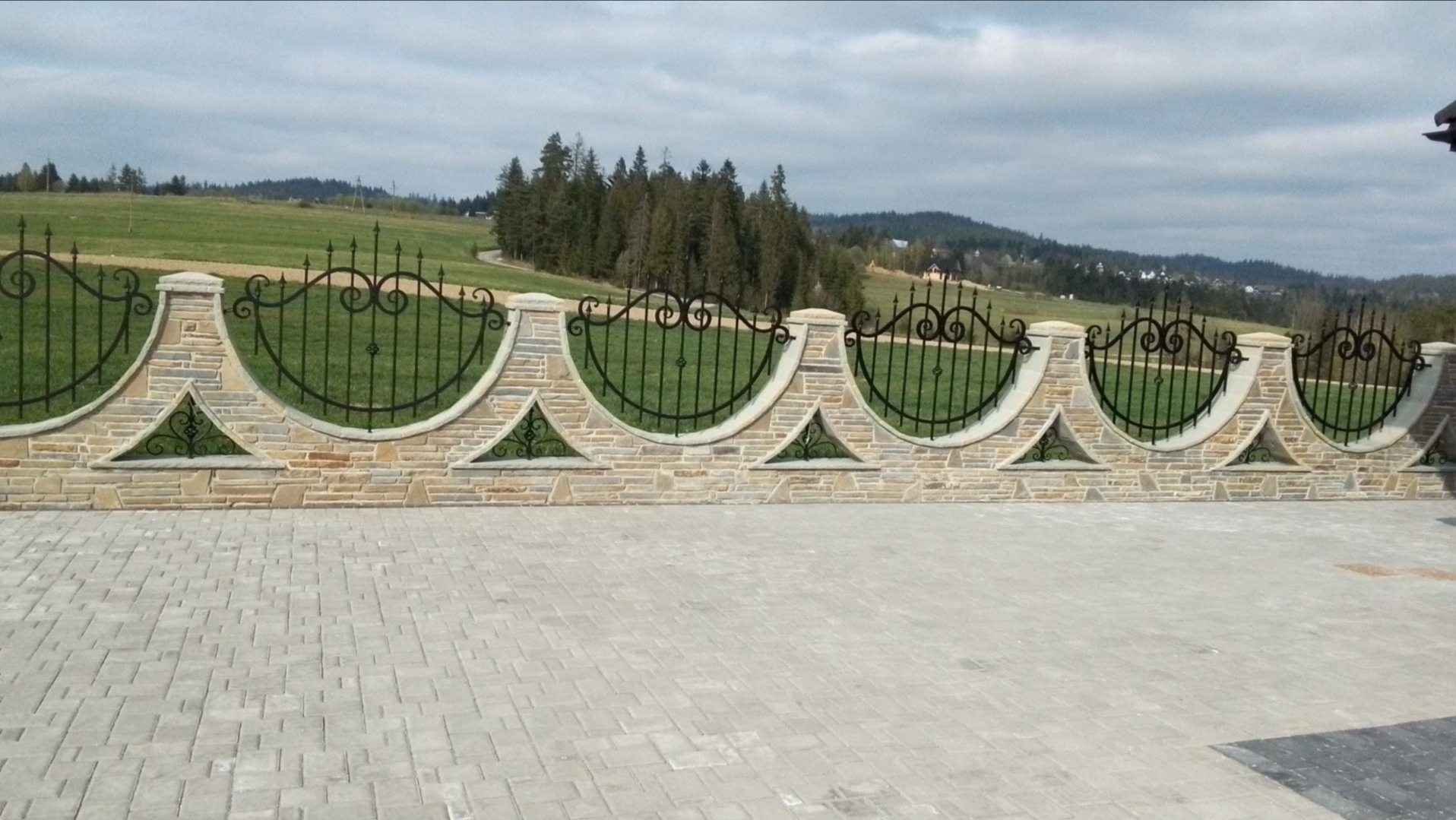 Kamienny murek z kutymi elementami ogrodzenia, widok na zielone wzgórza w tle, oraz ułożona kostka brukowa na podjeździe.