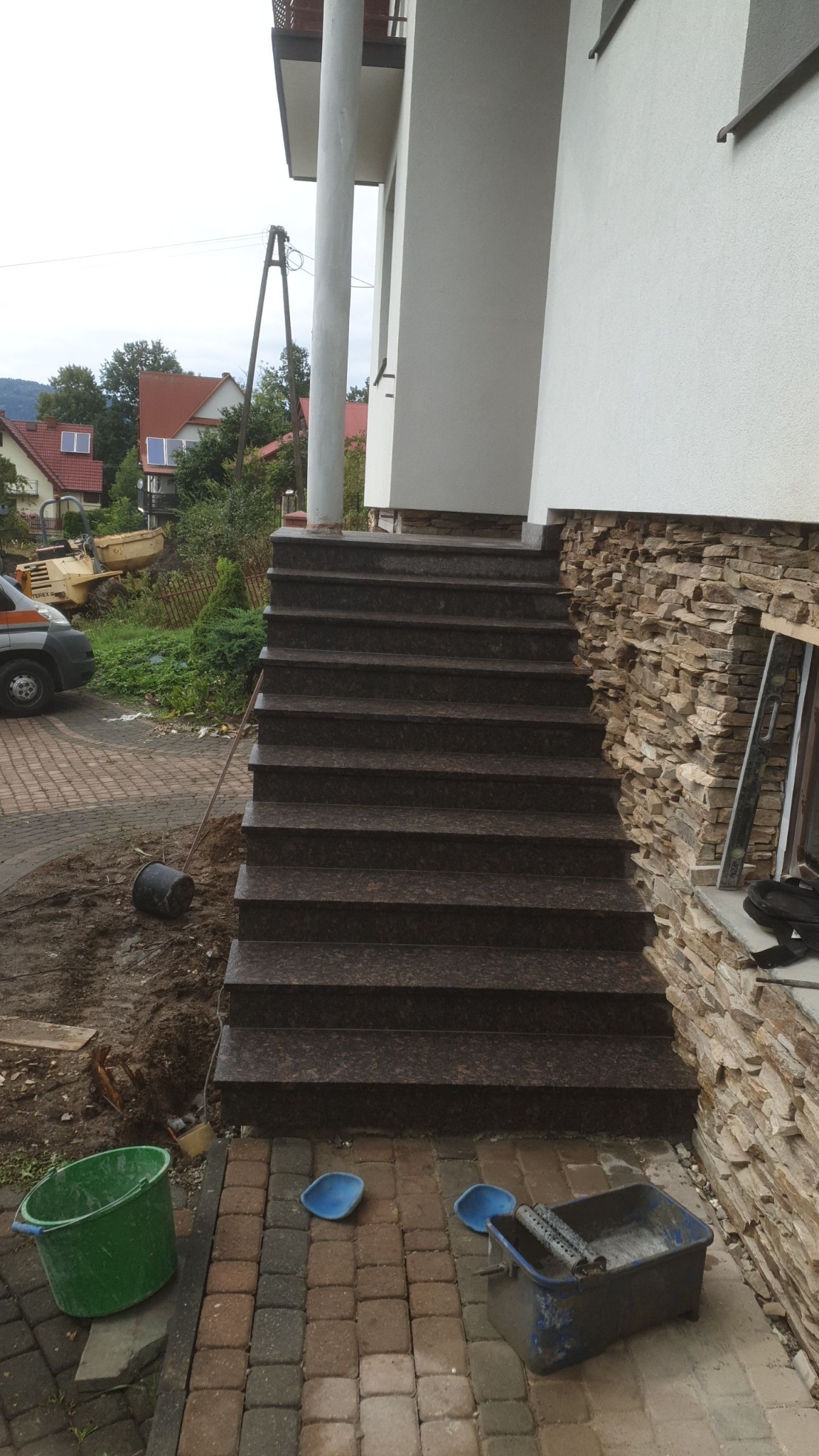 Granitowe schody zewnętrzne prowadzące do domu z elewacją z kamienia naturalnego, widoczne narzędzia i pozostałości po pracach budowlanych.