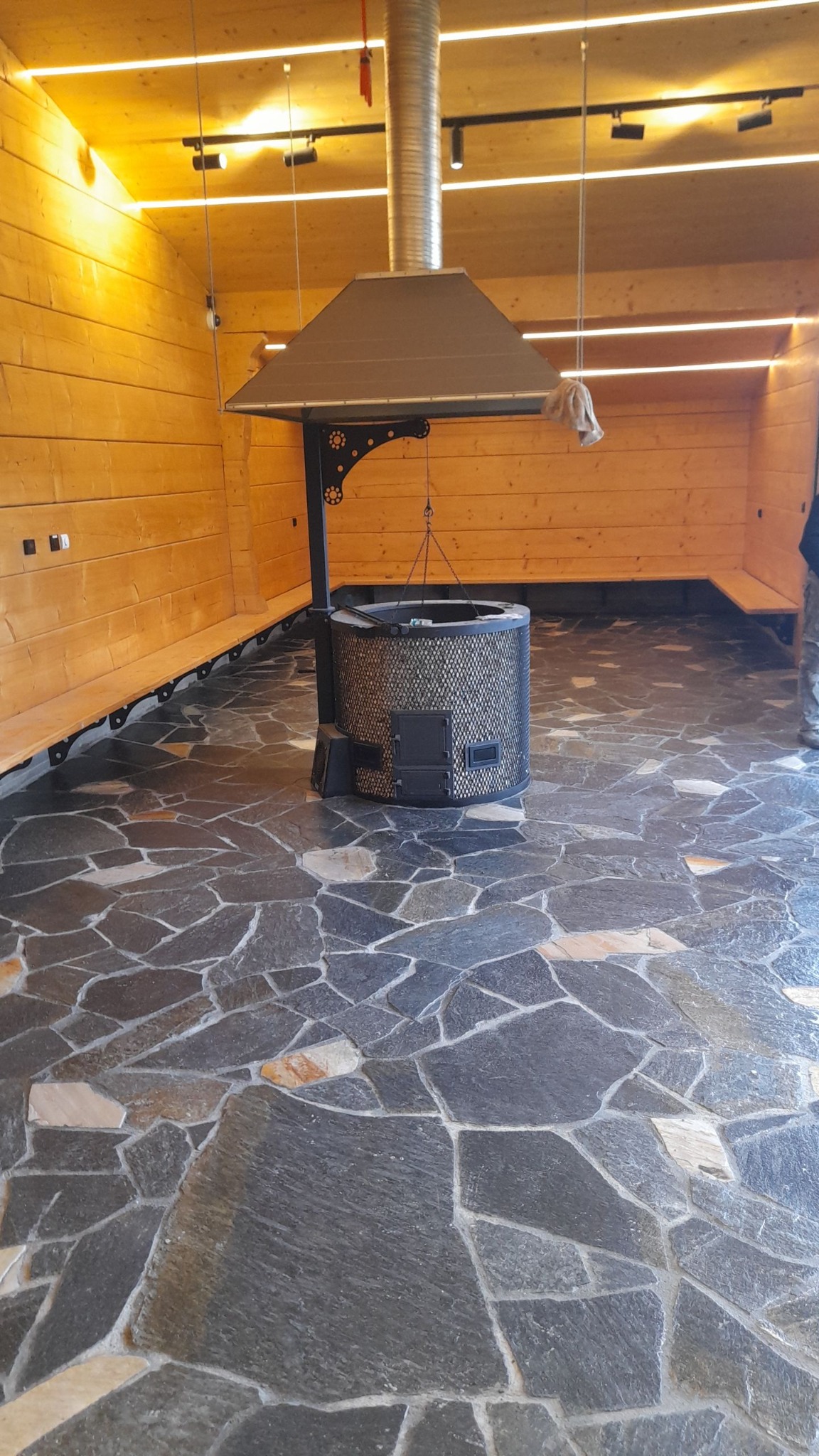 Kamienna posadzka z nieregularnych płyt w altanie z drewnianą zabudową i centralnie umieszczonym paleniskiem z kominem.