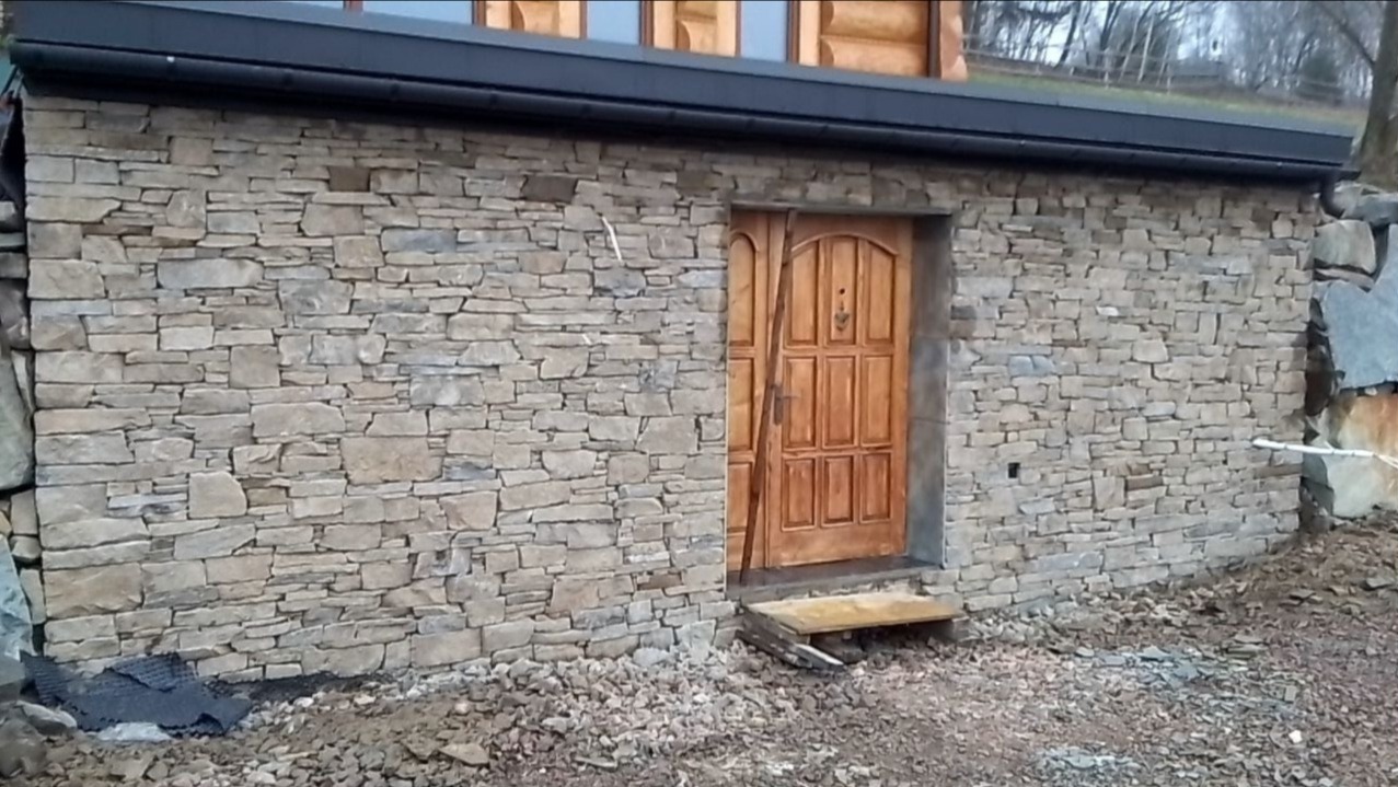 Ściana budynku obłożona nieregularnym kamieniem z drewnianymi drzwiami i prowizorycznym stopniem wejściowym.