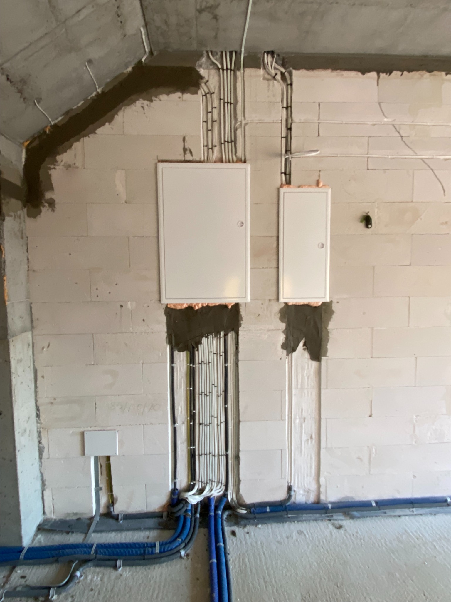 Surowa ściana z betonu komórkowego z dwoma białymi skrzynkami elektrycznymi, widoczne kable w peszlach i rurkach, niedokończone obróbki murarskie wokół przewodów.
