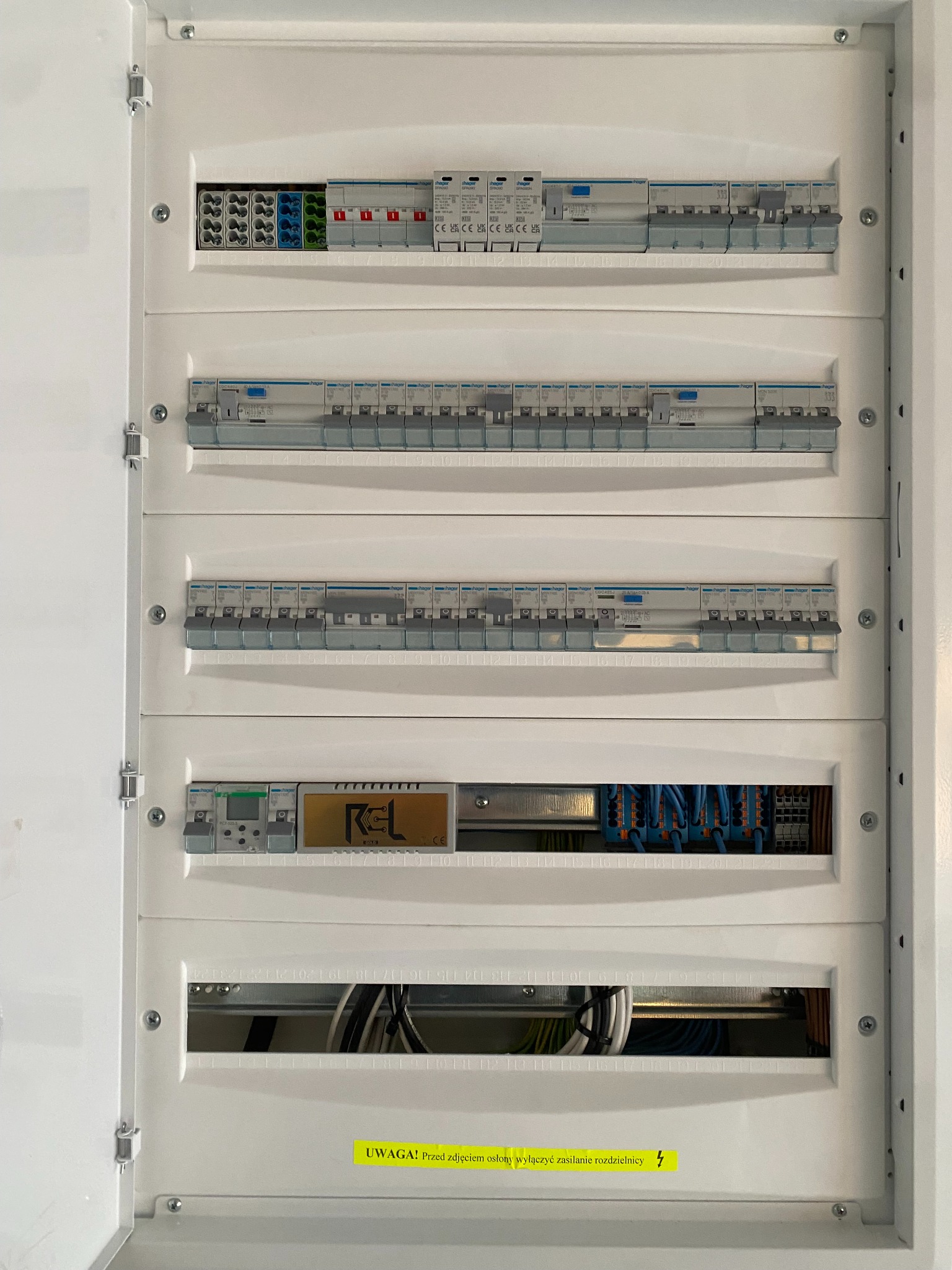 Otwarta, biała skrzynka rozdzielcza z zainstalowanymi bezpiecznikami, wyłącznikami różnicowoprądowymi i przewodami; widoczny napis ostrzegawczy na żółtej naklejce.