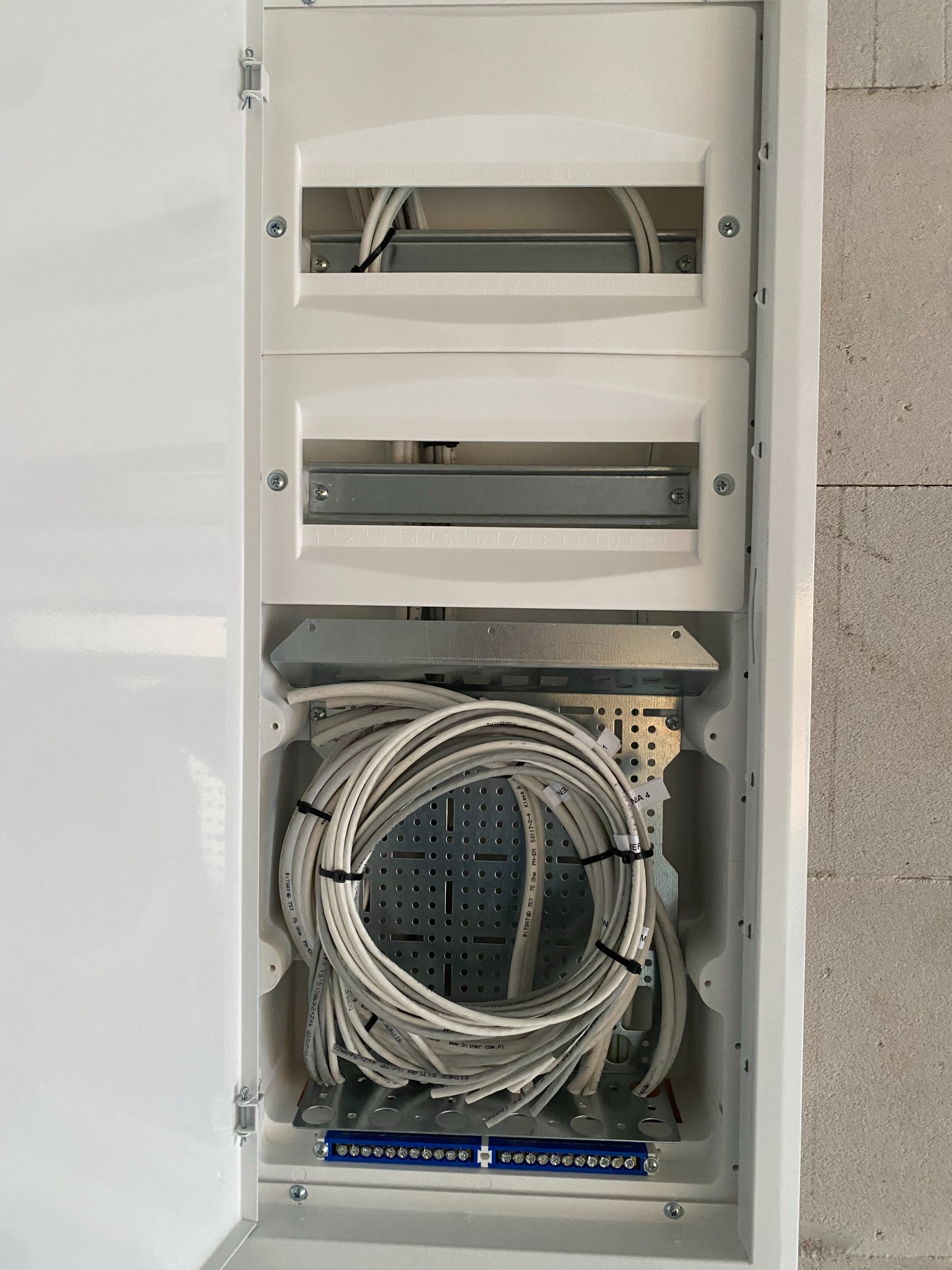 Otwarta skrzynka rozdzielcza z widocznymi modułami, metalowymi szynami montażowymi i zwiniętym kablem telekomunikacyjnym zabezpieczonym czarnymi opaskami, umieszczona na tle surowej ściany z betonu...