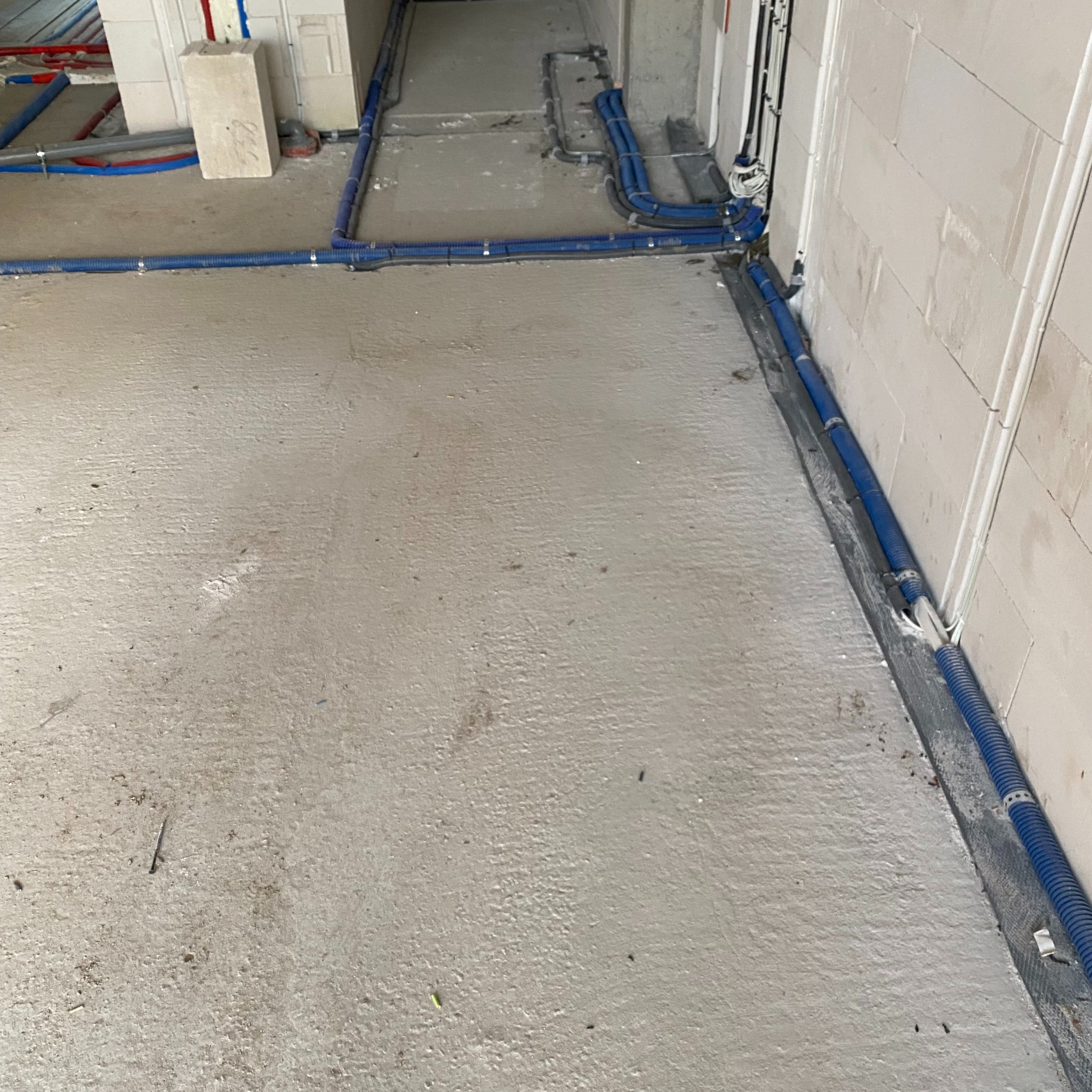 Niebieskie rury instalacyjne prowadzone po posadzce w surowym stanie, mocowane do ścian z białego betonu komórkowego, widoczne fragmenty innych instalacji.