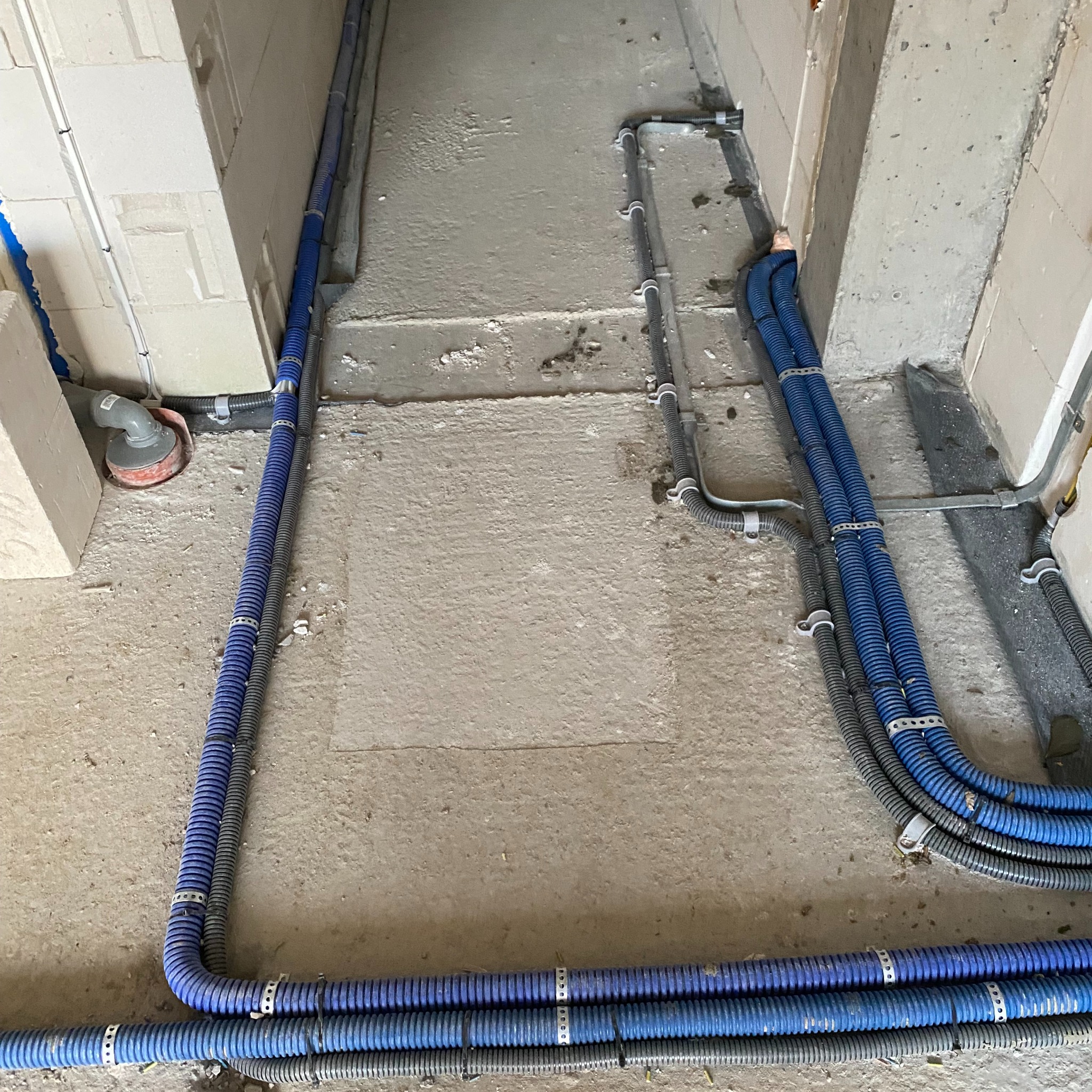 Instalacja elektryczna w trakcie montażu: niebieskie i szare rury karbowane prowadzone wzdłuż ścian z betonu komórkowego, mocowane metalowymi uchwytami.