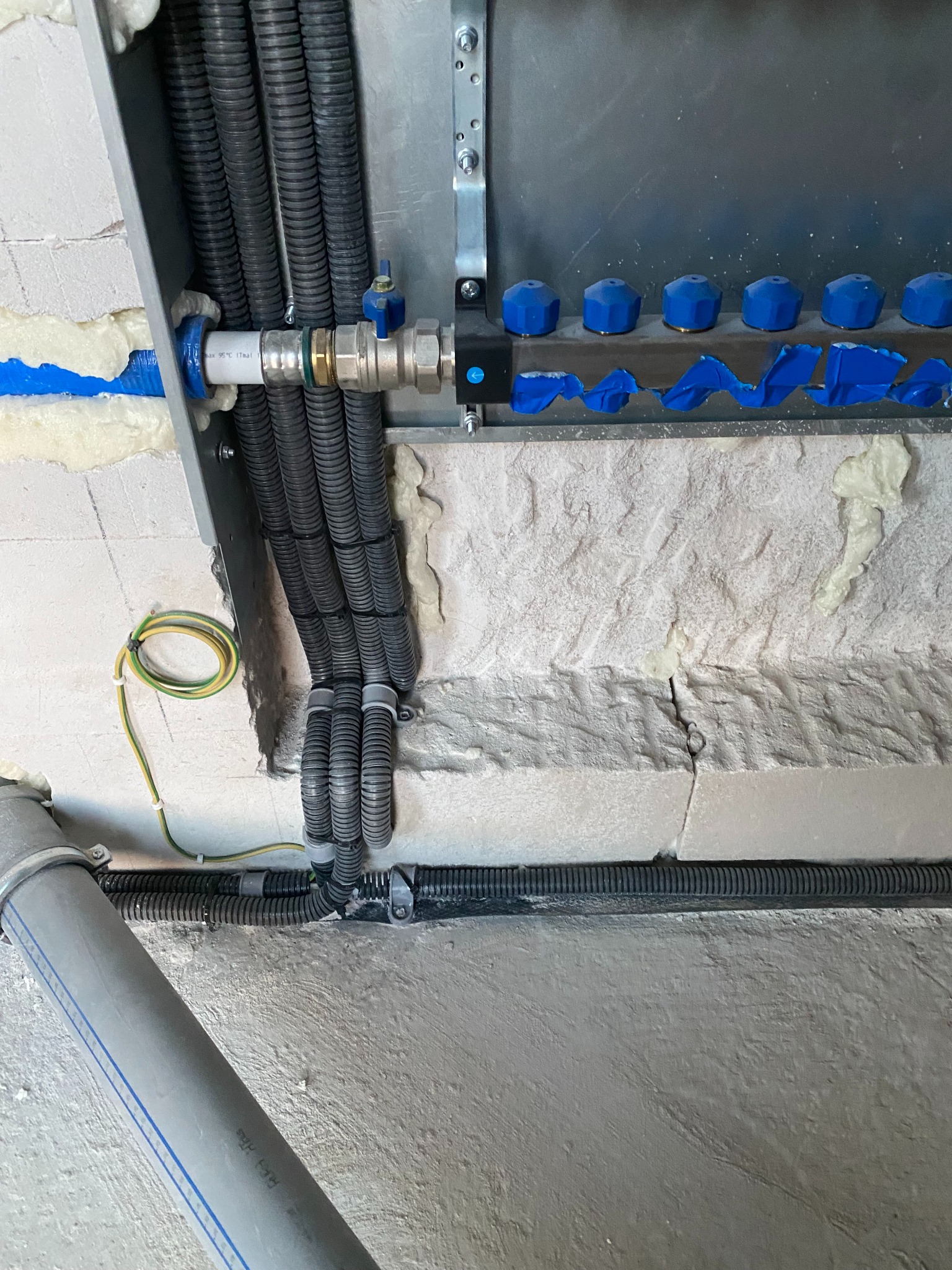 Rozdzielacz instalacji podłogowej z niebieskimi zaworami, widoczne liczne kable w peszlach oraz rura odpływowa w szarej ścianie z cegły, ocieplenie pianką montażową.