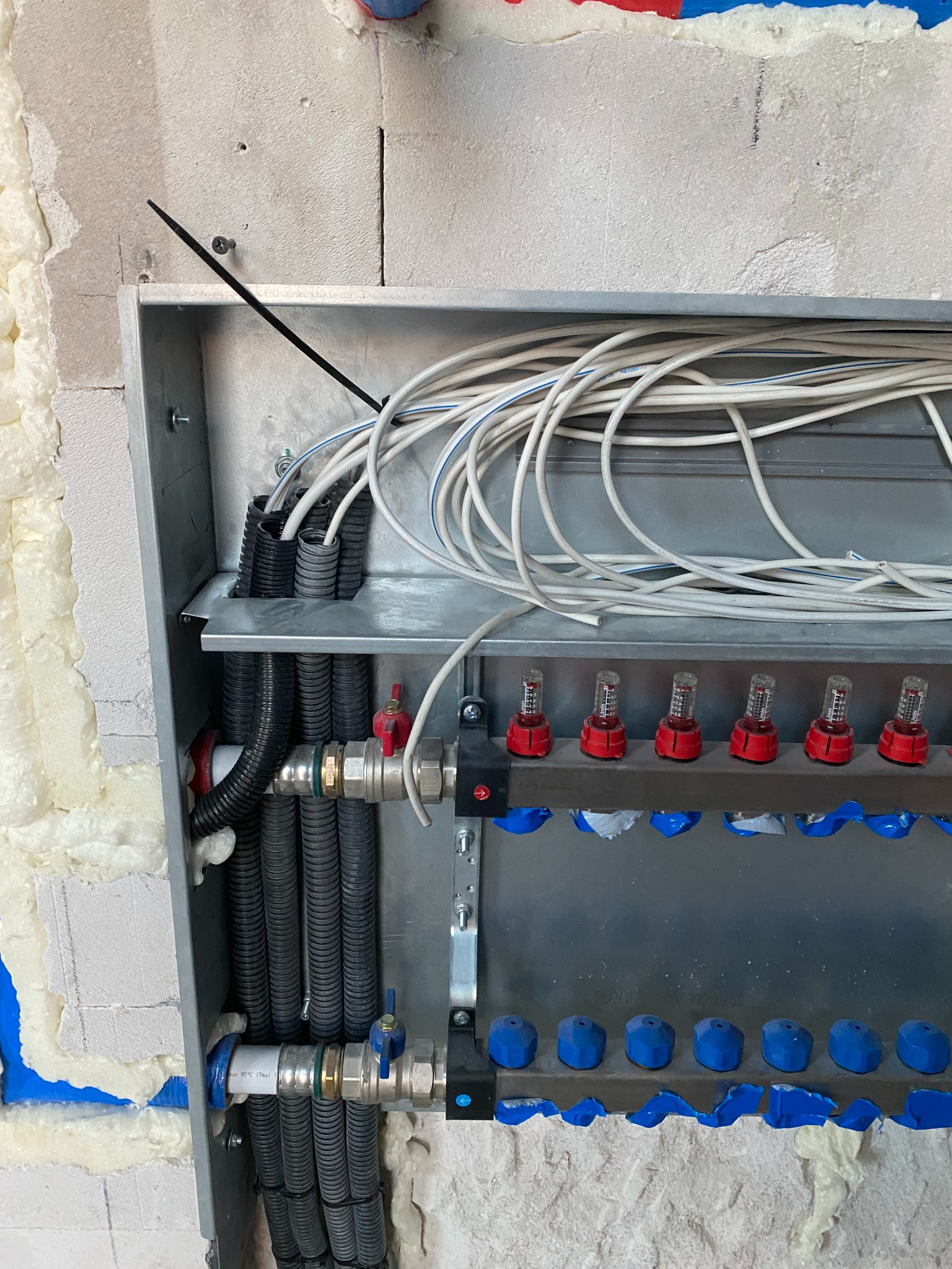 Rozdzielacz instalacji grzewczej w metalowej obudowie, widoczne rury, zawory, przepływomierze z czerwonymi elementami, białe przewody elektryczne i czarne peszle, montaż na ścianie z izolacją...