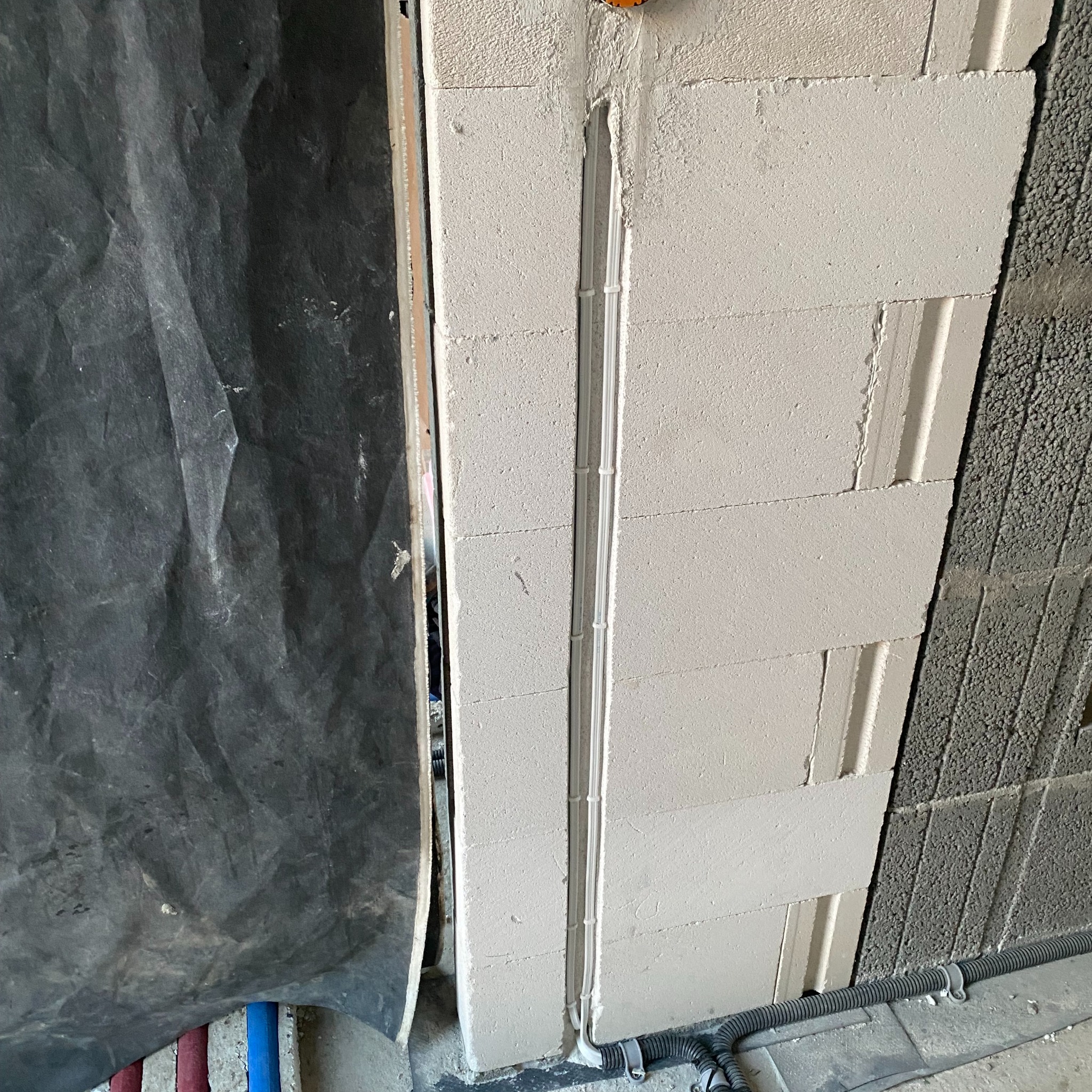 Białe kable elektryczne prowadzone w bruździe w ścianie z betonu komórkowego, zabezpieczone peszlem przy podłodze, obok ściana z szarego bloczka betonowego i folia ochronna.